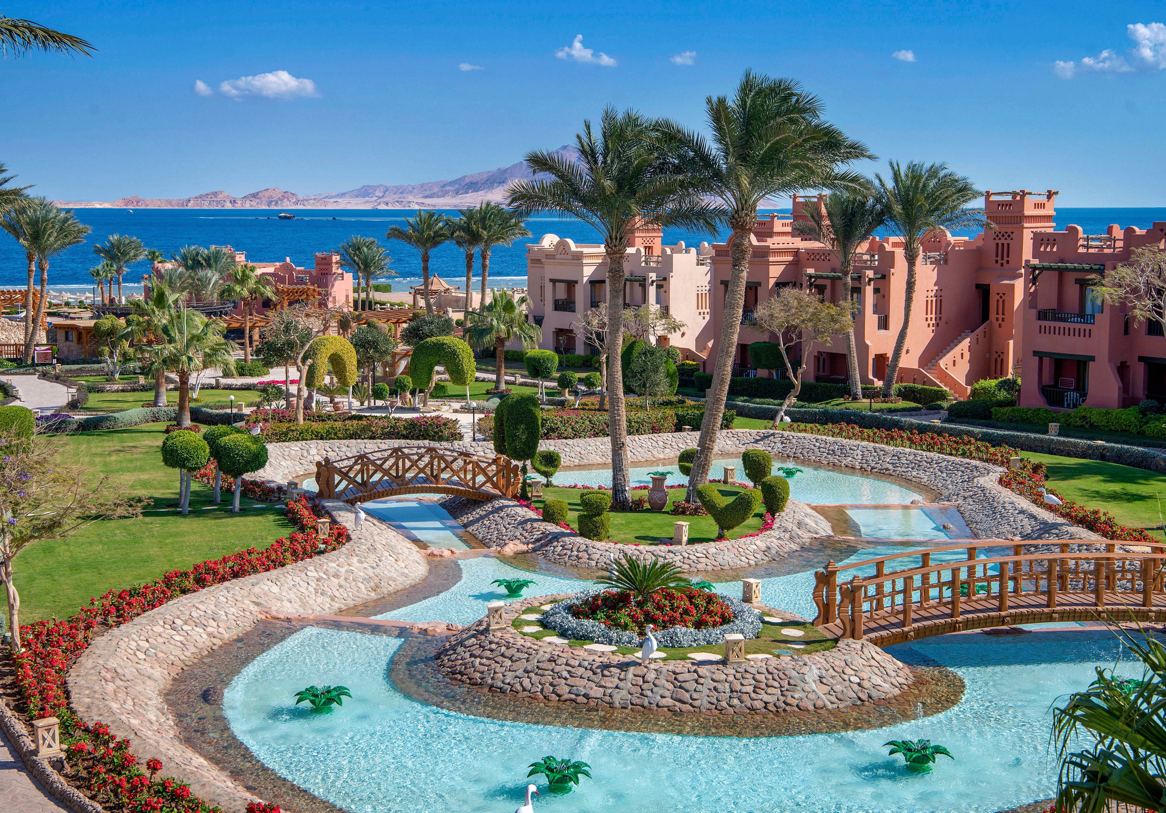 A picturesque luxury resort featuring manicured gardens, flowing water features, and wooden bridges. The setting overlooks a serene ocean with distant mountains under a clear blue sky. Palm trees and vibrant flowers enhance the tropical ambiance.