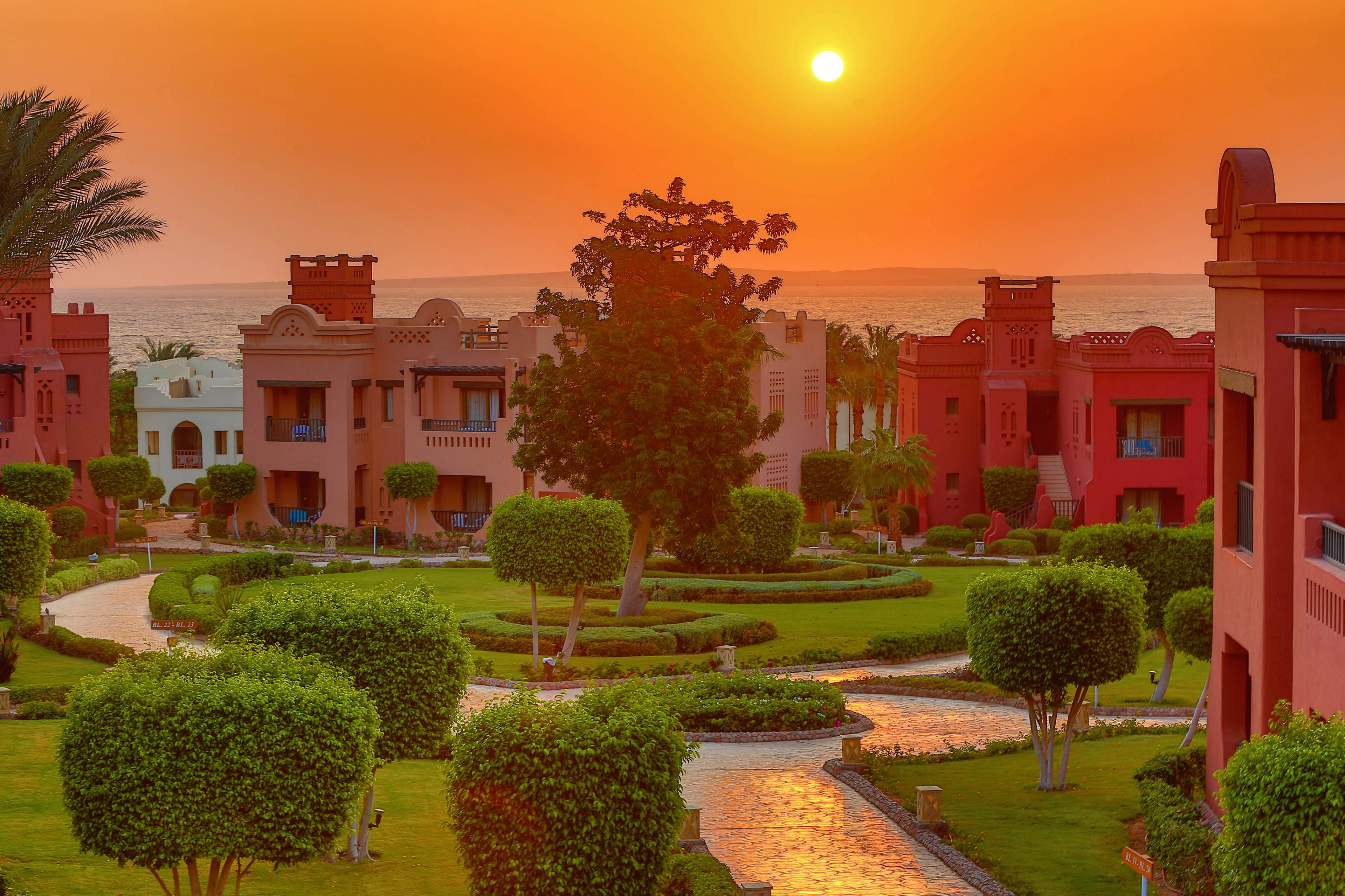 A vibrant sunset illuminates a resort with red-toned buildings and lush green gardens. The scene features a winding pathway surrounded by manicured trees and shrubs, leading to the ocean in the background. The warm orange hues of the sky create a serene and inviting atmosphere.