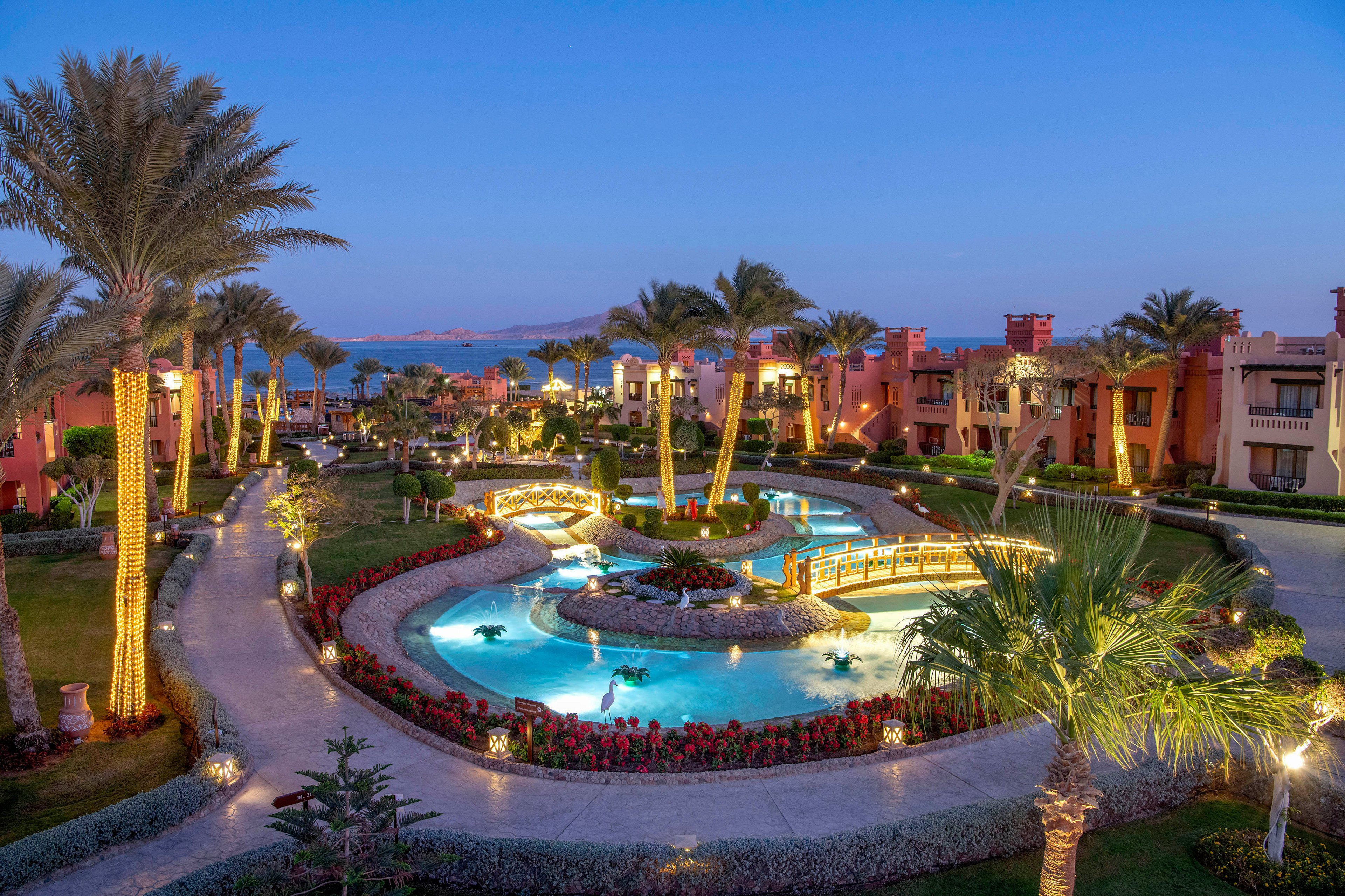 A luxurious resort features a beautifully illuminated pool surrounded by lush greenery and palm trees at dusk. The setting includes elegant pathways, vibrant landscaping, and a serene ambiance. The distant view of the sea and softly lit buildings enhances the tranquil atmosphere.