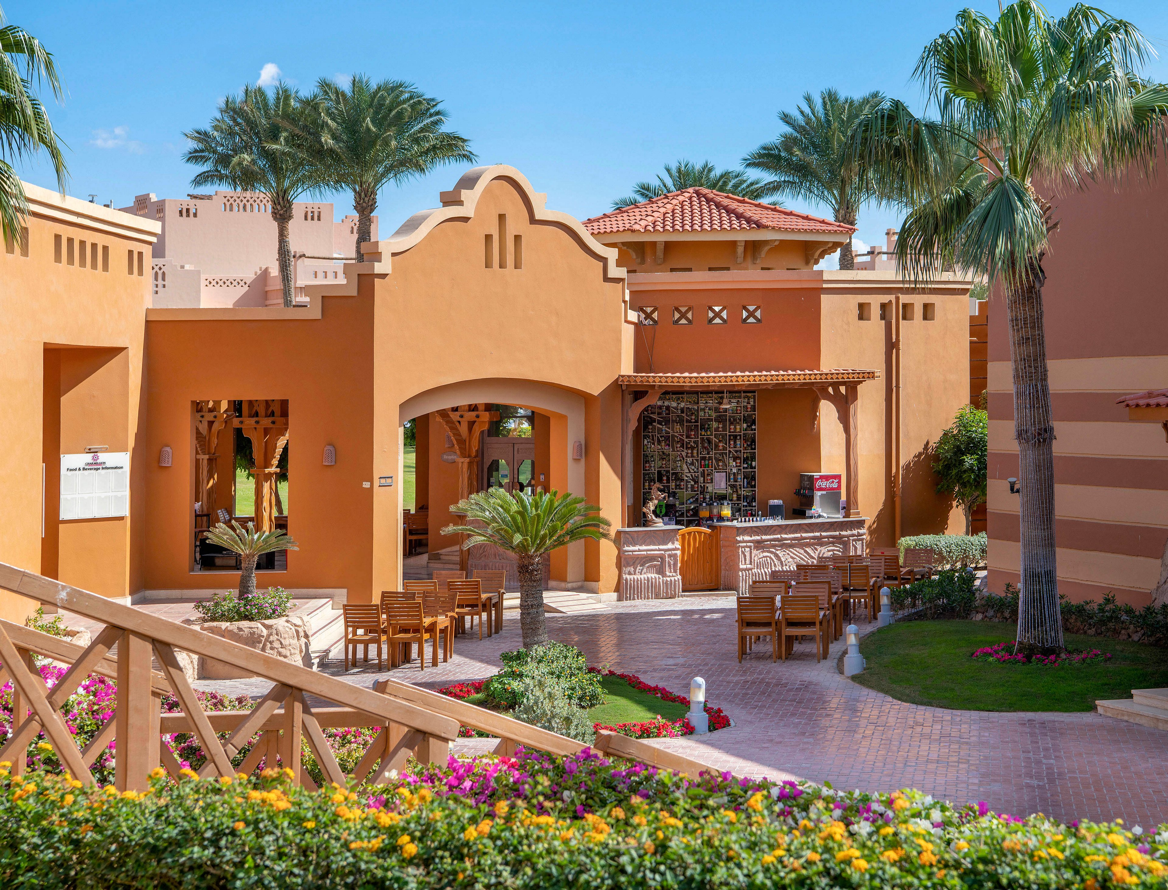 A charming outdoor cafe set amidst vibrant gardens and Mediterranean-style architecture. The setting features warm terracotta tones, lush palm trees, and colorful flowers. Wooden tables and chairs are arranged in a welcoming courtyard under a clear blue sky.
