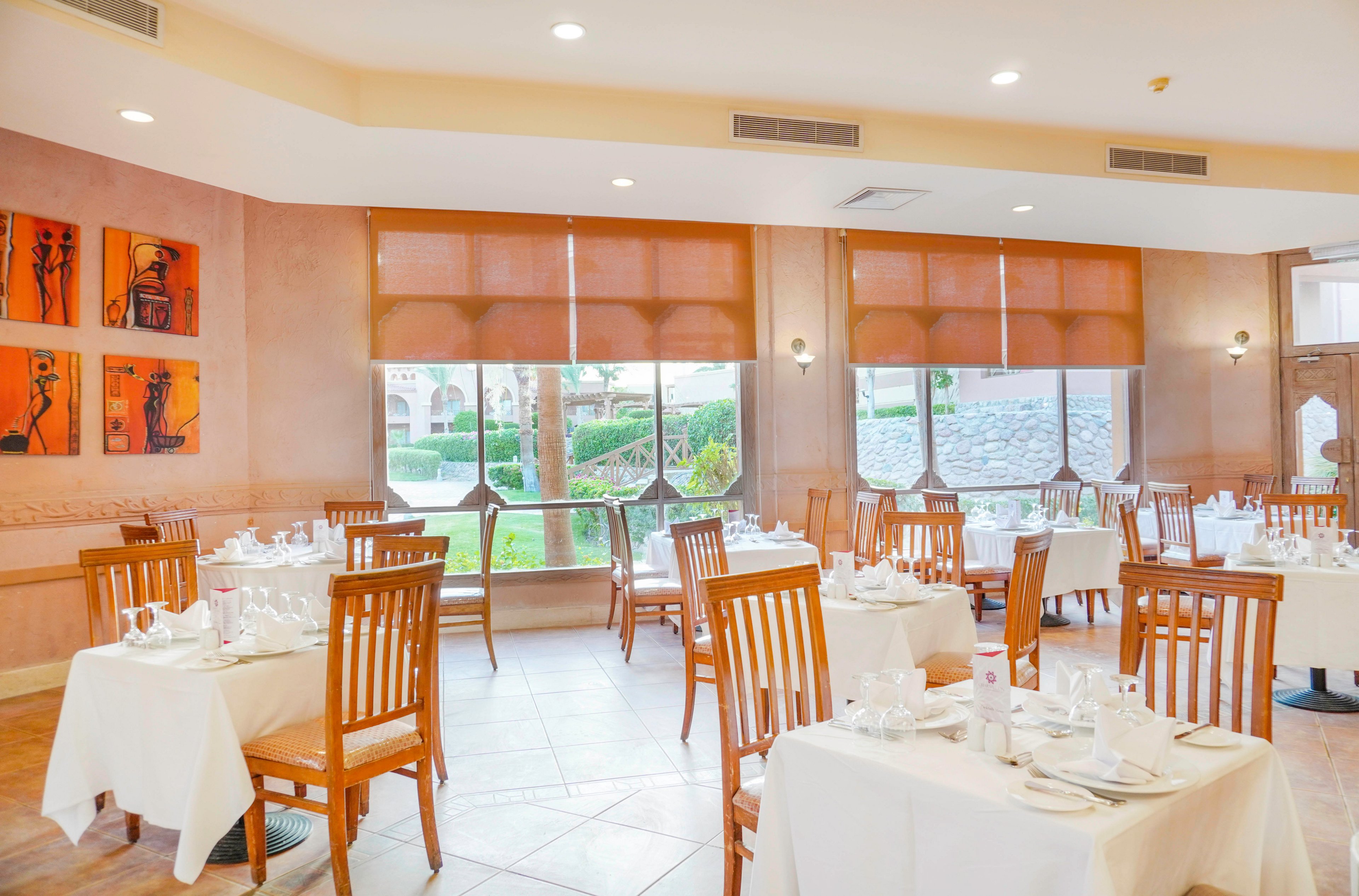 A well-lit restaurant dining area featuring neatly arranged wooden chairs and tables with white tablecloths and place settings. Large windows allow natural light to flood the space, offering a view of a landscaped garden. Warm-toned walls are adorned with modern artwork, creating a welcoming ambiance.