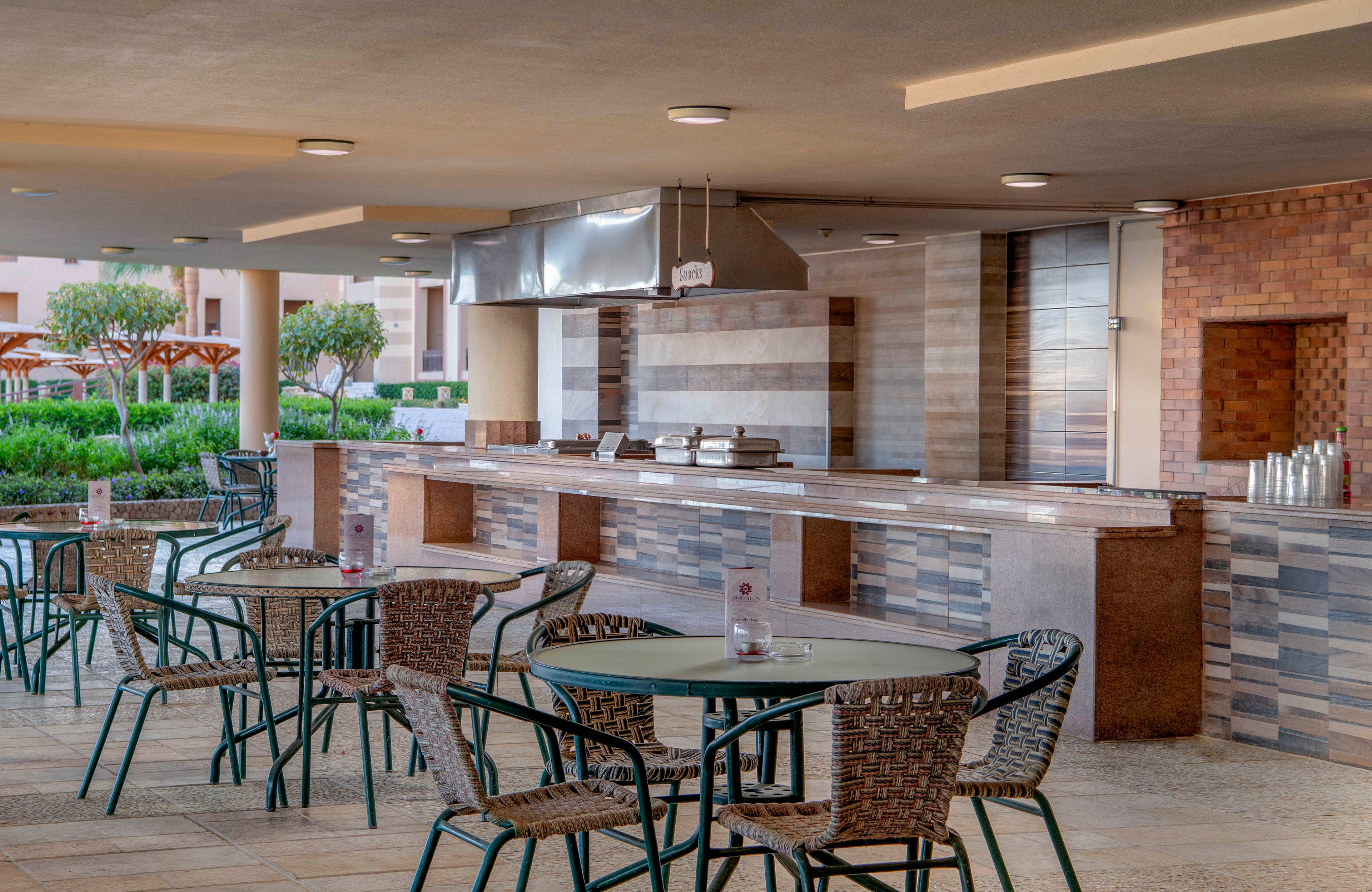 An open-air dining area featuring a modern design with tiled walls and a stainless steel cooking station. The space includes multiple tables and chairs arranged neatly under a covered patio. Greenery and resort-style umbrellas are visible in the background, creating a relaxed atmosphere.