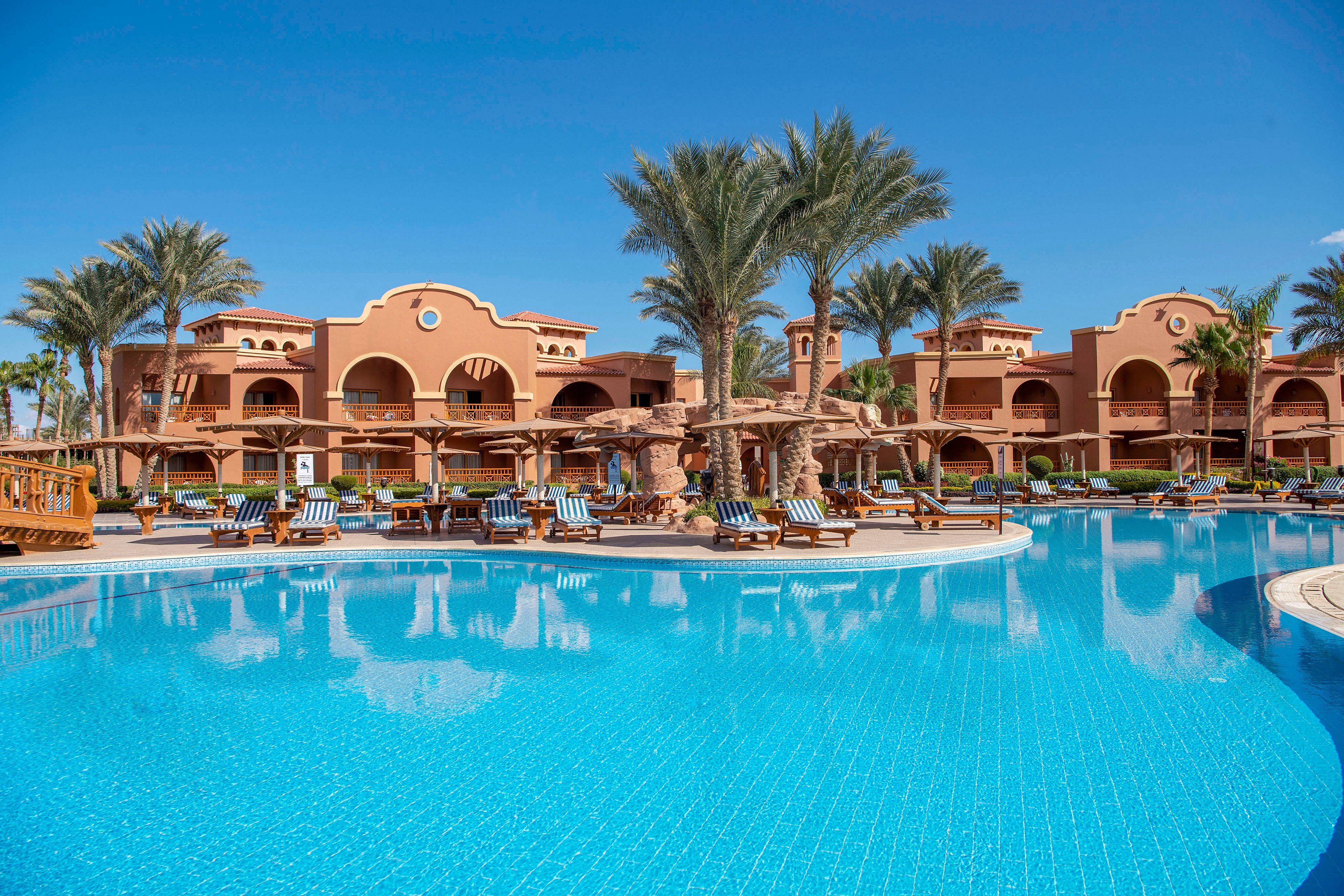 A luxurious resort featuring a large, clear blue swimming pool surrounded by palm trees and sun loungers. The architecture showcases warm terracotta tones with arches and balconies. The setting is sunny and inviting, perfect for relaxation and leisure activities.