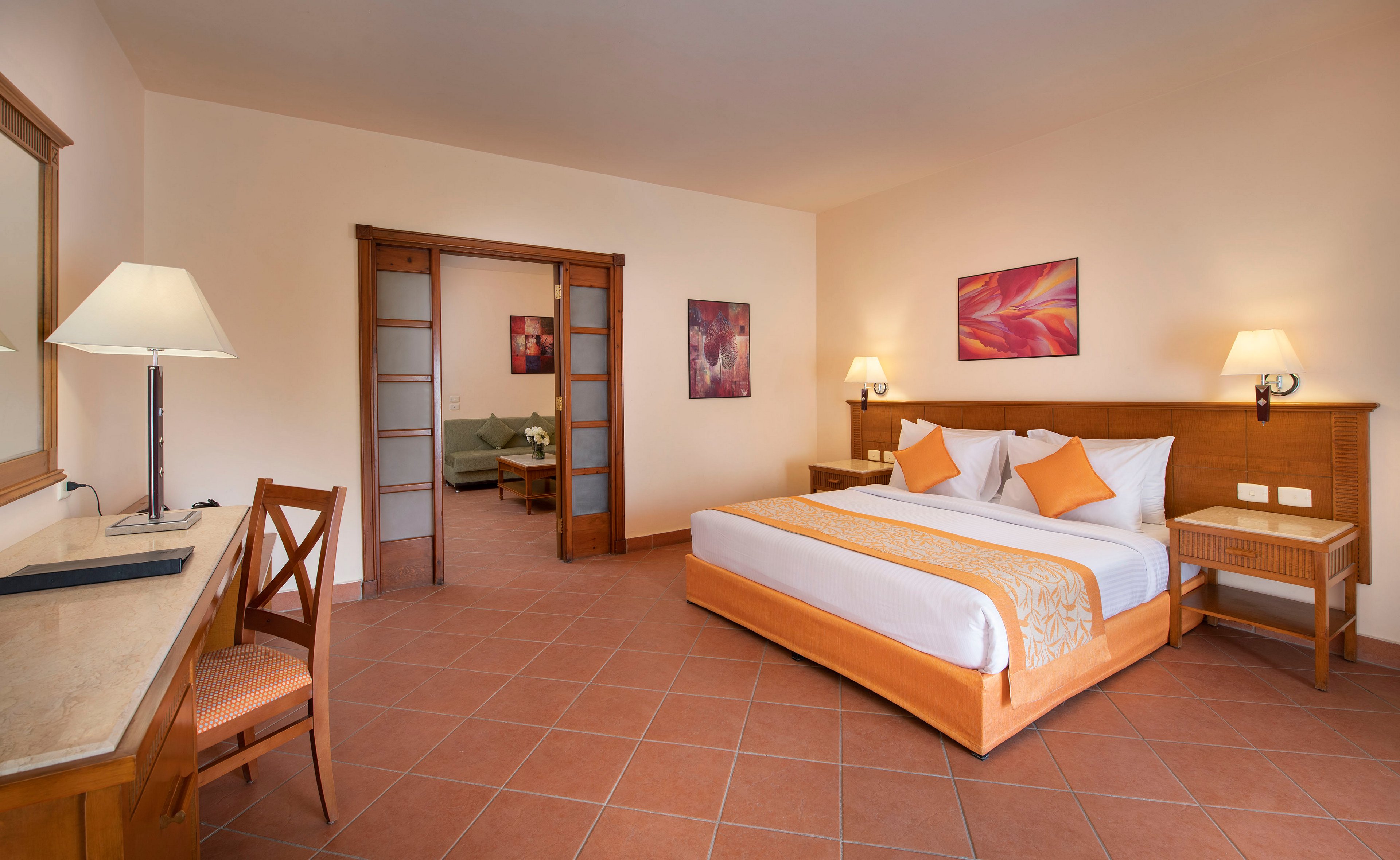 A spacious hotel room featuring a neatly made bed with orange accents, a wooden desk, and a seating area visible through sliding doors. The room is well-lit with table lamps and natural light. Warm tones dominate the decor, complemented by tiled flooring and abstract wall art.