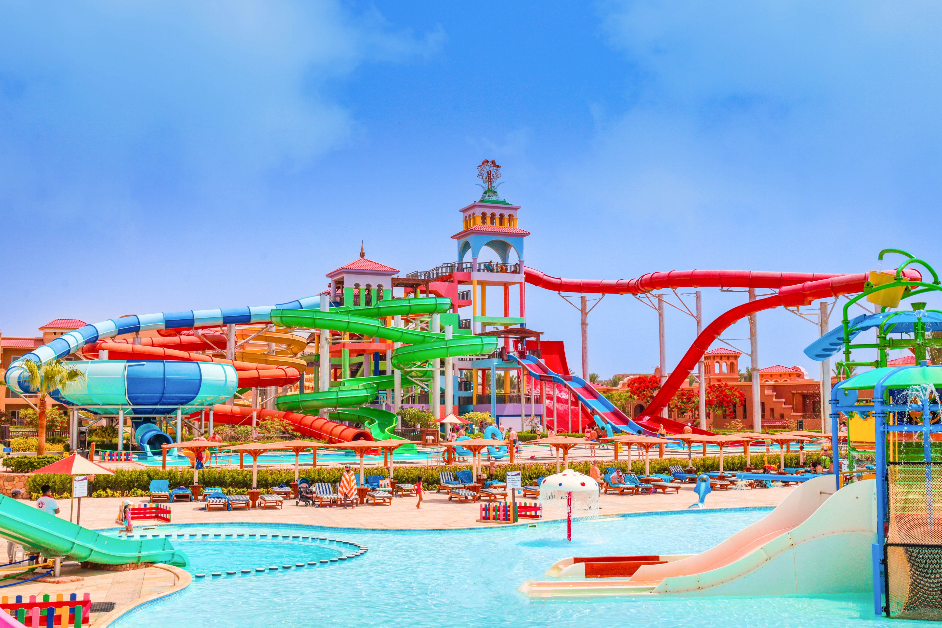 A vibrant water park featuring multiple colorful slides and large pools under a bright blue sky. The setting includes various attractions such as winding tubes, tall towers, and splash zones. The area is designed for leisure and family fun, with seating and umbrellas visible around the pools.