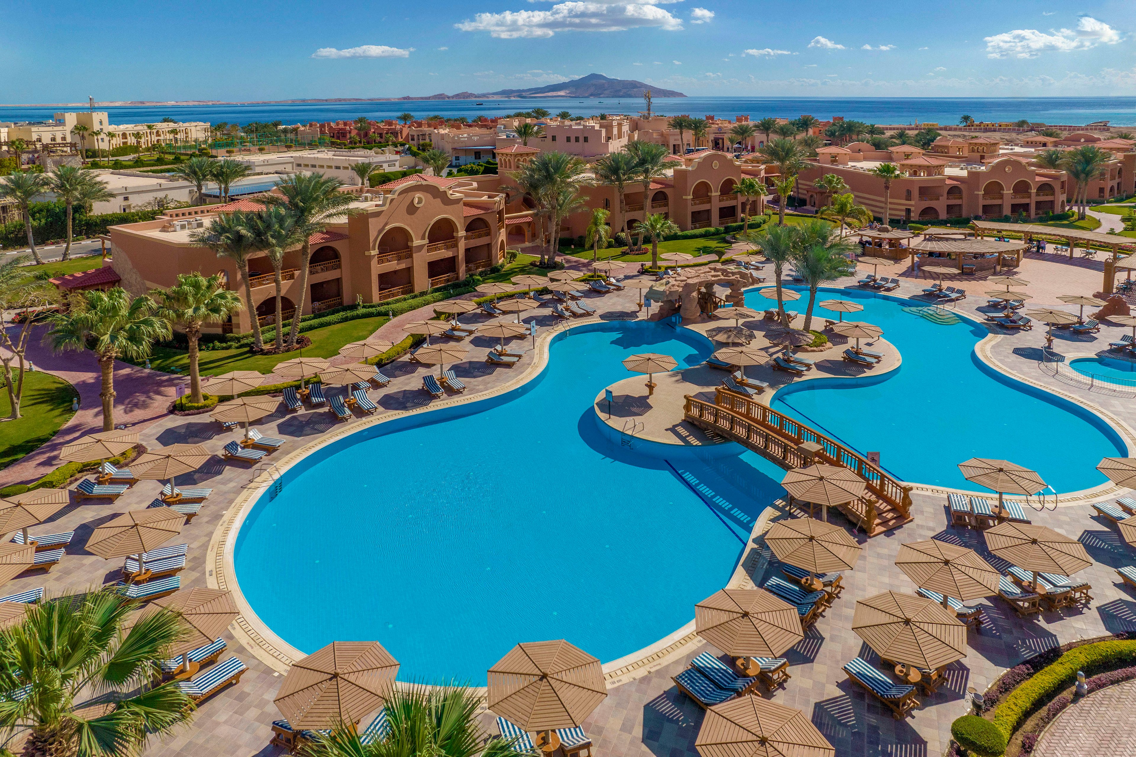 A large, uniquely shaped swimming pool surrounded by sun loungers and umbrellas in a luxury resort setting. The resort features Mediterranean-style architecture with palm trees and lush landscaping. In the background, a serene ocean view and distant mountains enhance the tranquil atmosphere. The bright blue sky complements the vibrant colors of the pool and surroundings.