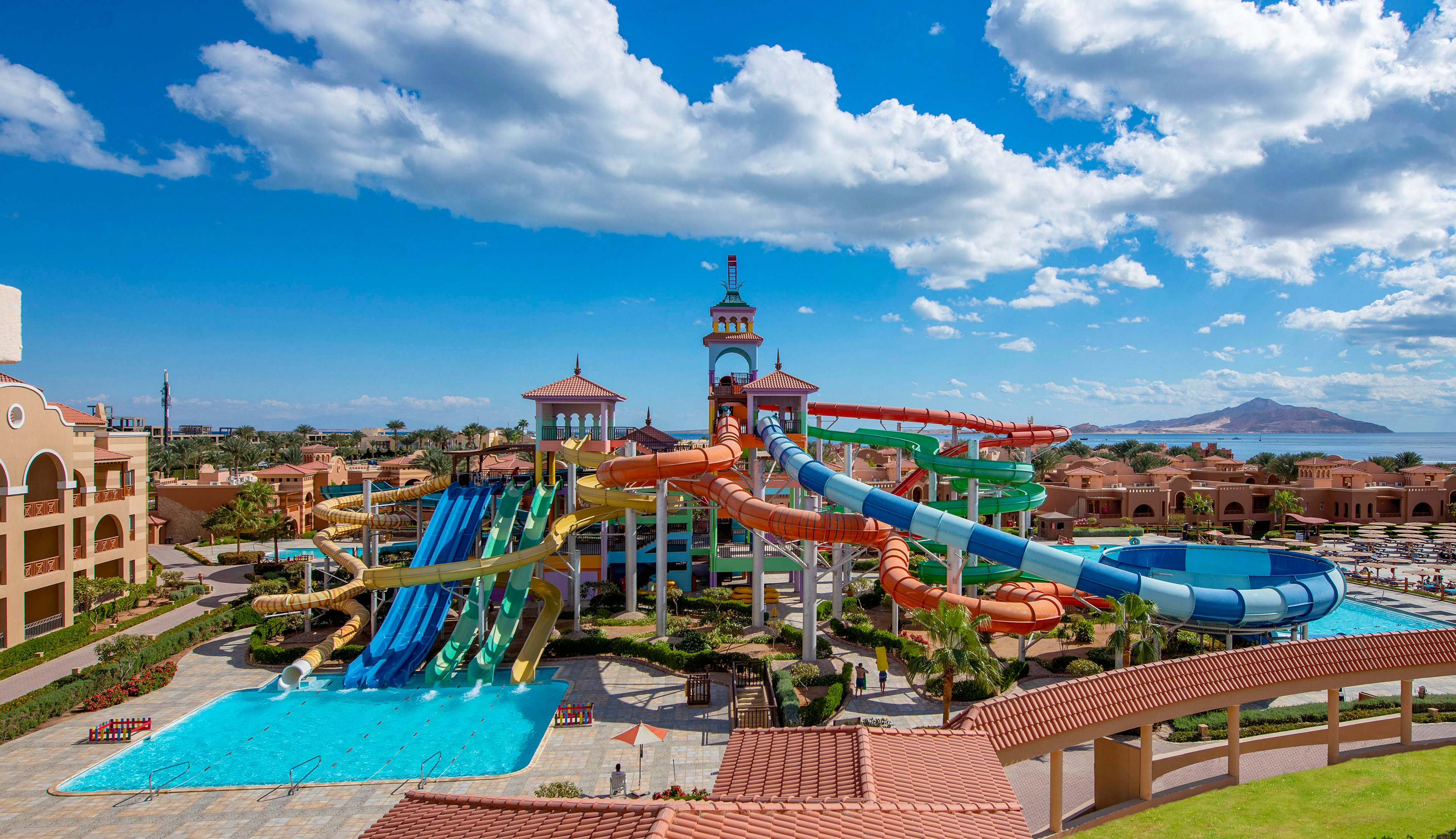 A vibrant water park featuring multiple colorful water slides and a large pool under a bright blue sky. The setting includes a tower structure and surrounding resort buildings, with scenic mountains visible in the background. The area is well-maintained with pathways and greenery, creating a lively and inviting atmosphere.