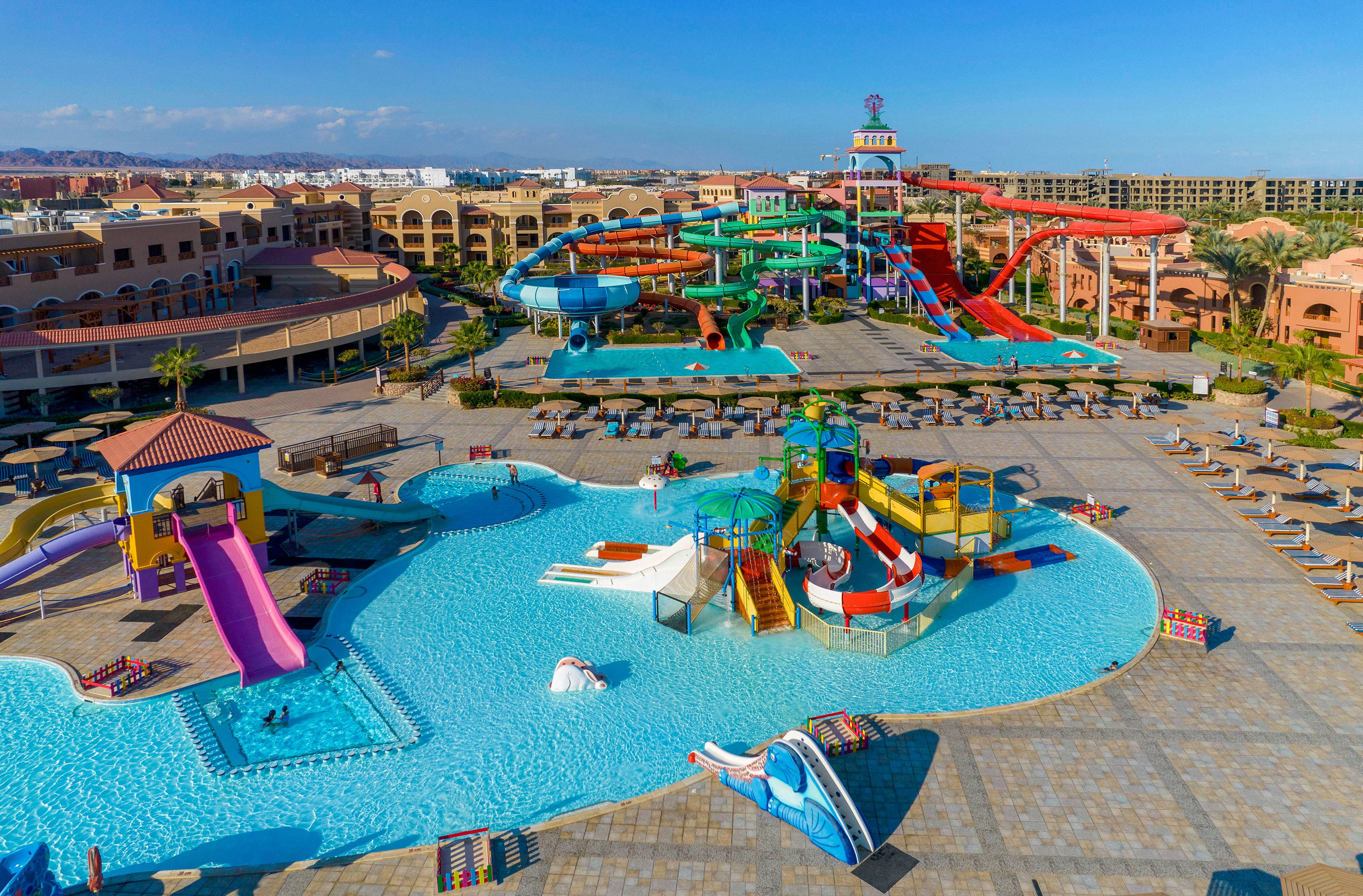A vibrant water park featuring multiple colorful slides, a large pool, and a children's play area. The setting includes sun loungers and umbrellas, surrounded by resort buildings under a clear blue sky. The park offers a fun and lively atmosphere for families and visitors.