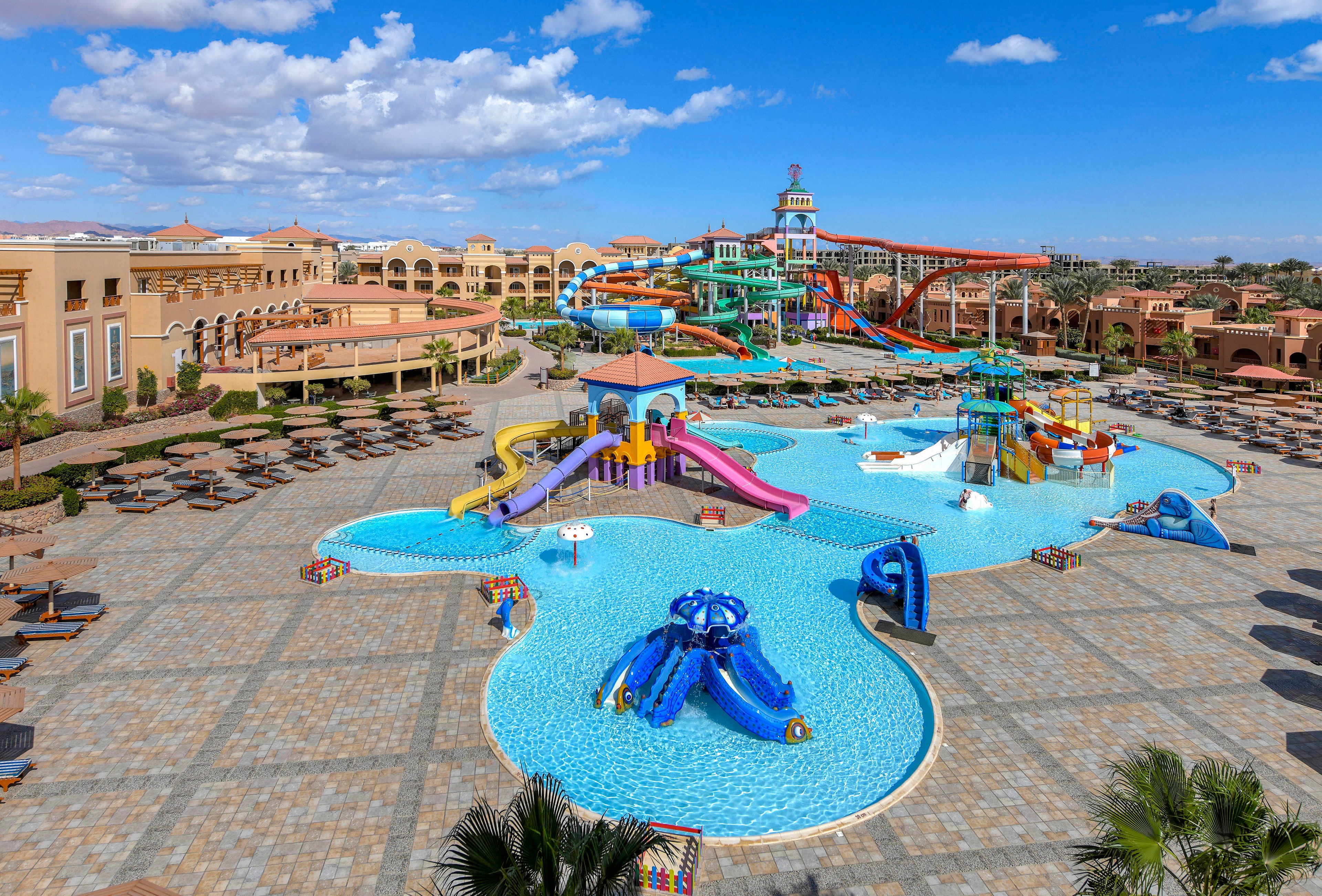 A sprawling water park featuring multiple colorful slides, pools, and play areas. The setting includes a sunny day with clear skies, surrounded by resort-style buildings and lounge chairs. The vibrant design and spacious layout make it ideal for family recreation and leisure activities.