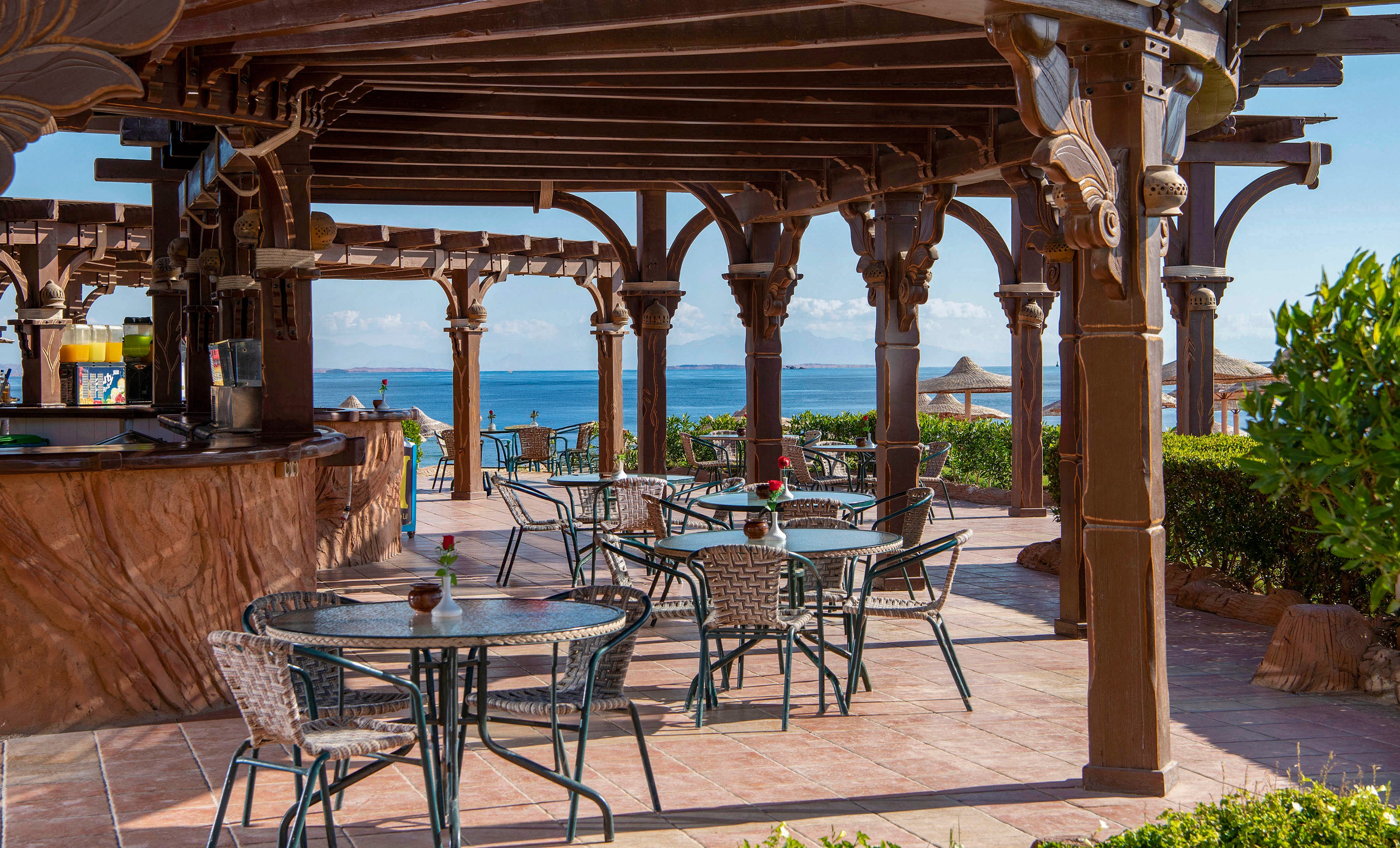 An open-air café with intricately designed wooden pergolas overlooking a serene seaside view. The setting features empty tables and chairs on a tiled patio, surrounded by lush greenery. The ocean and straw umbrellas in the background add to the tranquil atmosphere.
