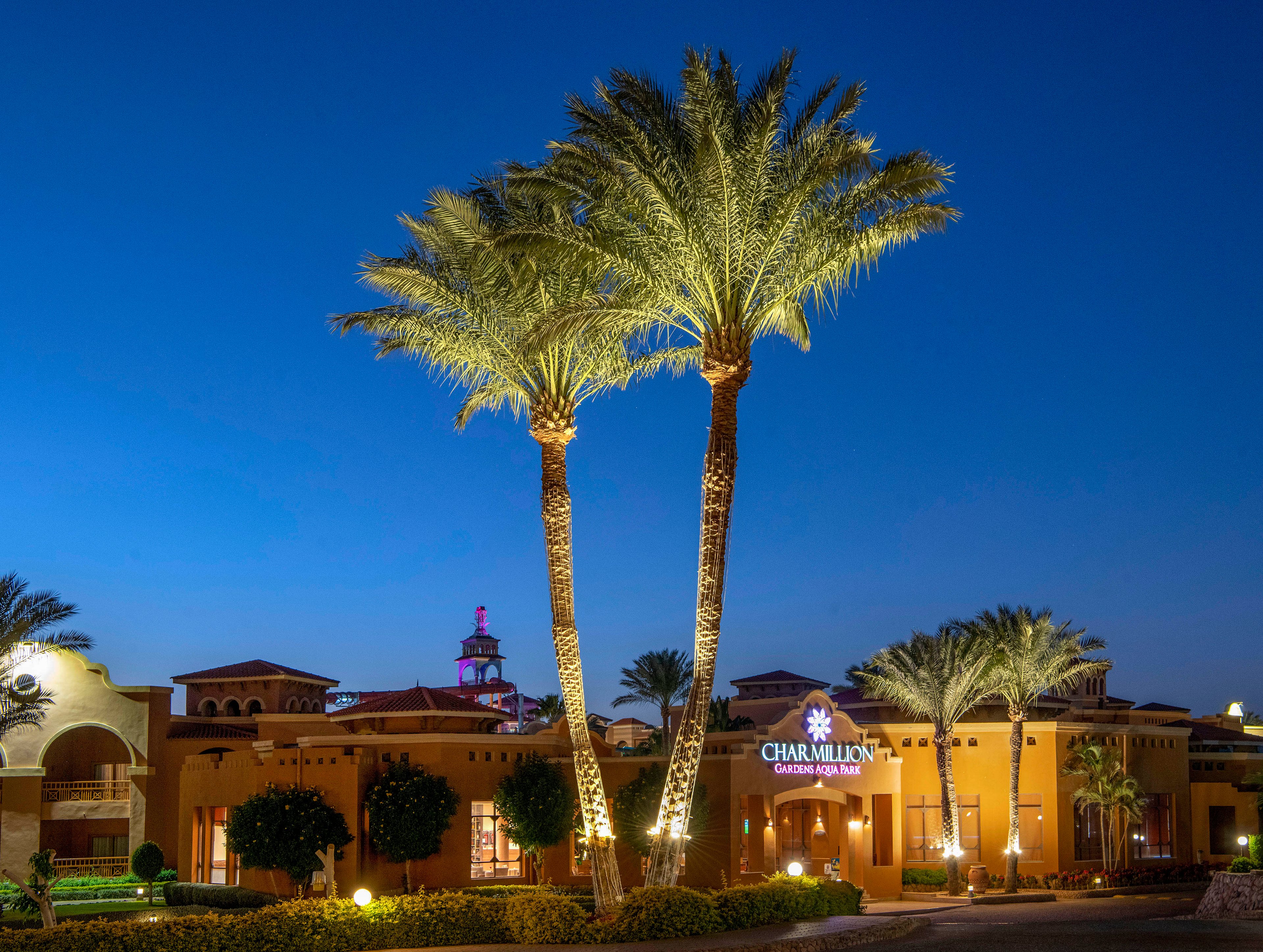 Illuminated palm trees stand tall in front of a resort entrance during twilight. The building features warm lighting and Mediterranean-style architecture. A visible sign reads 'Charmillion Gardens Aqua Park,' adding a sense of location and purpose to the scene.