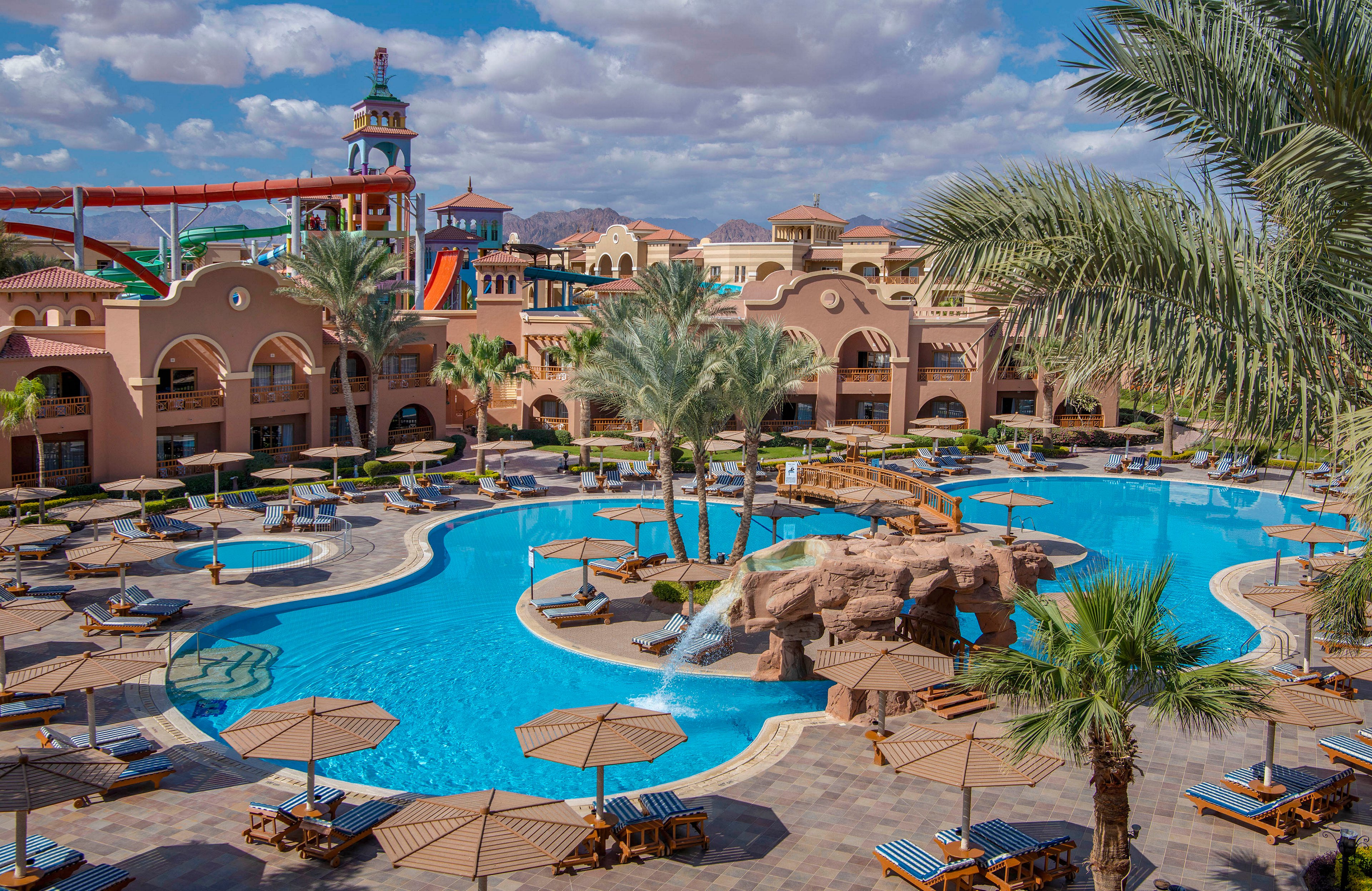 A luxurious resort featuring a large outdoor pool surrounded by lounge chairs and umbrellas. The setting includes palm trees, a rock fountain, and vibrant water slides in the background. The architecture showcases warm tones and Mediterranean-style buildings under a partly cloudy sky.