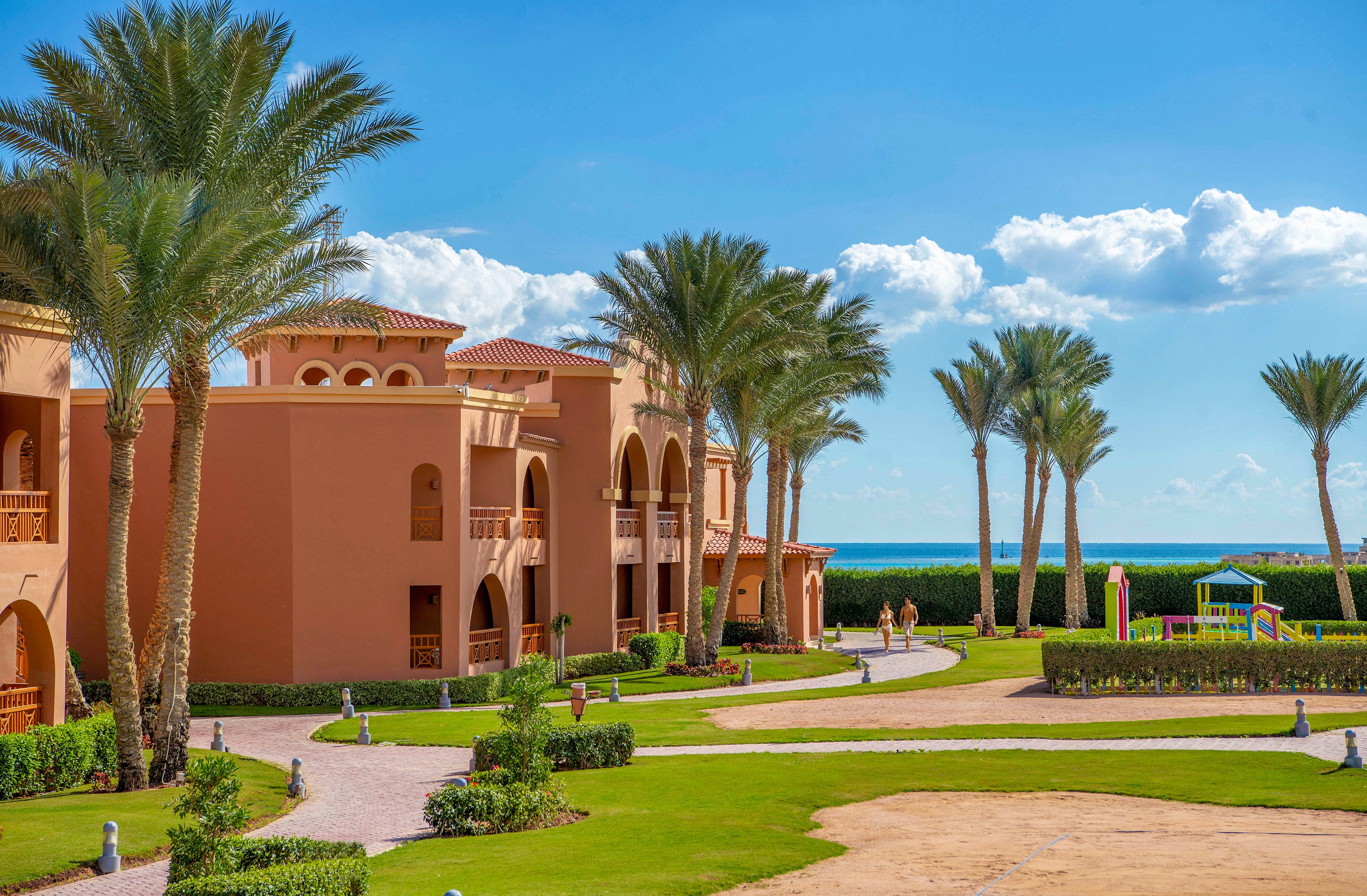 A scenic resort featuring terracotta-colored buildings surrounded by lush green lawns and tall palm trees. The setting includes a clear blue sky and a view of the ocean in the background. A small playground is visible, adding a family-friendly touch to the serene environment.
