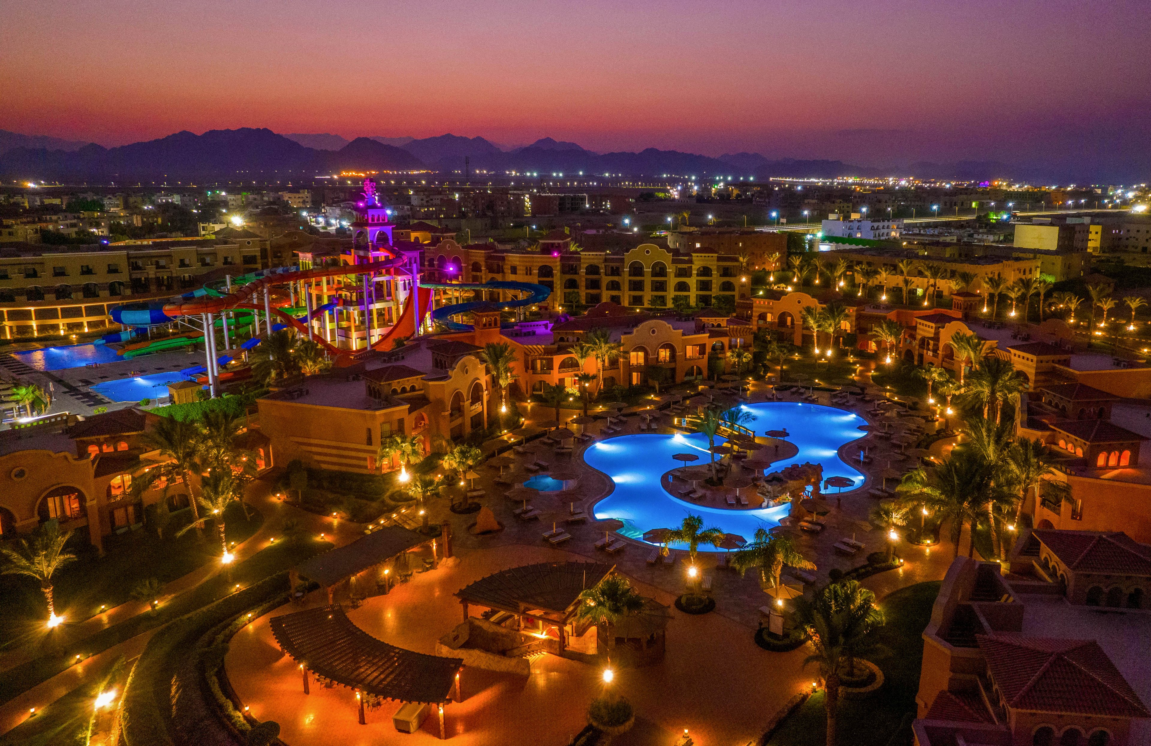 A stunning aerial view of a resort illuminated at dusk, featuring multiple pools and colorful water slides. The setting is surrounded by lush palm trees and vibrant lighting, creating a lively and inviting atmosphere. The backdrop includes distant mountains and a softly lit cityscape under a purple and orange sky.