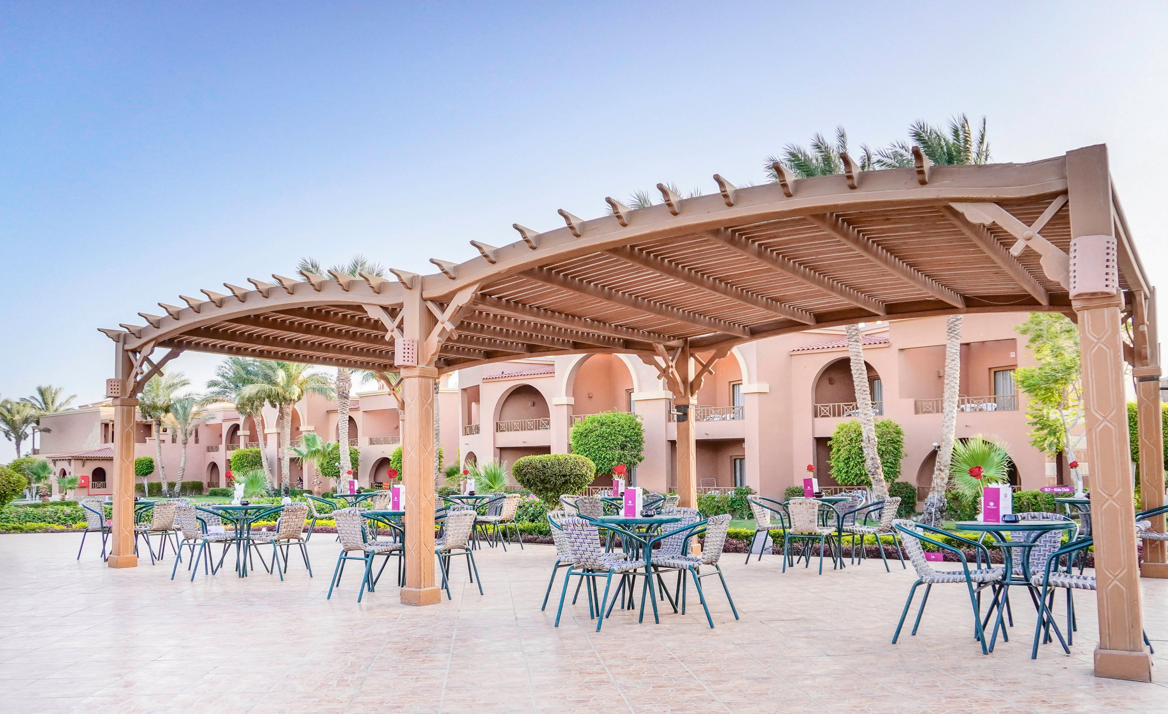 An outdoor patio area featuring a wooden pergola with multiple tables and chairs arranged for dining or relaxation. The setting is surrounded by lush greenery and palm trees, with a backdrop of peach-colored buildings. The clear blue sky adds to the serene and inviting atmosphere.