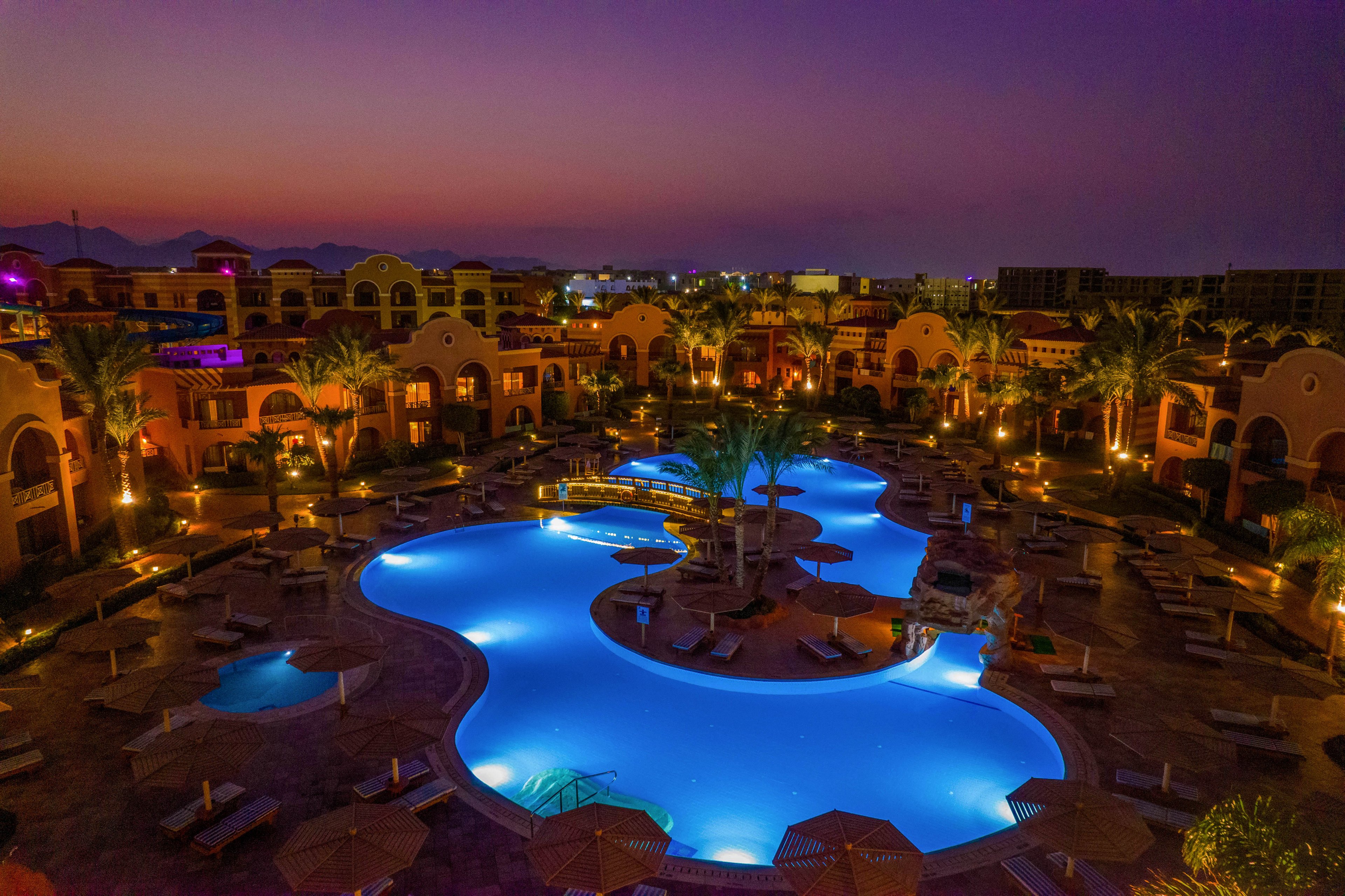 A beautifully illuminated resort pool with a unique shape is captured at dusk. The surrounding area features lounge chairs, palm trees, and warm lighting, creating a serene and luxurious ambiance. The sky transitions from deep purple to orange, enhancing the tranquil atmosphere.