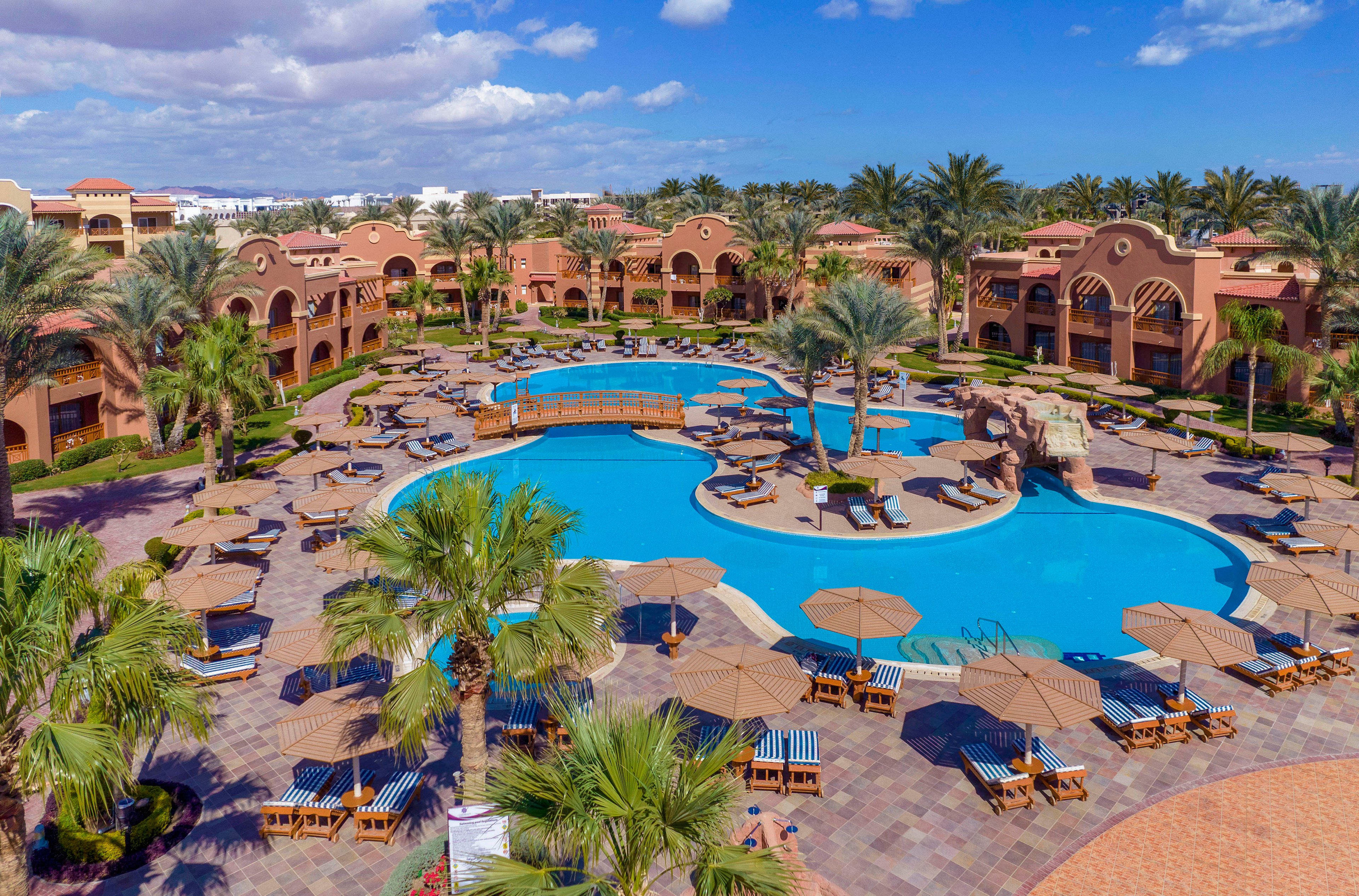 Aerial view of a luxury resort featuring a large, curving swimming pool surrounded by palm trees and sun loungers. The setting includes terracotta-colored buildings with arches and balconies, creating a Mediterranean-inspired atmosphere. The clear blue sky and lush greenery enhance the tranquil and inviting mood.
