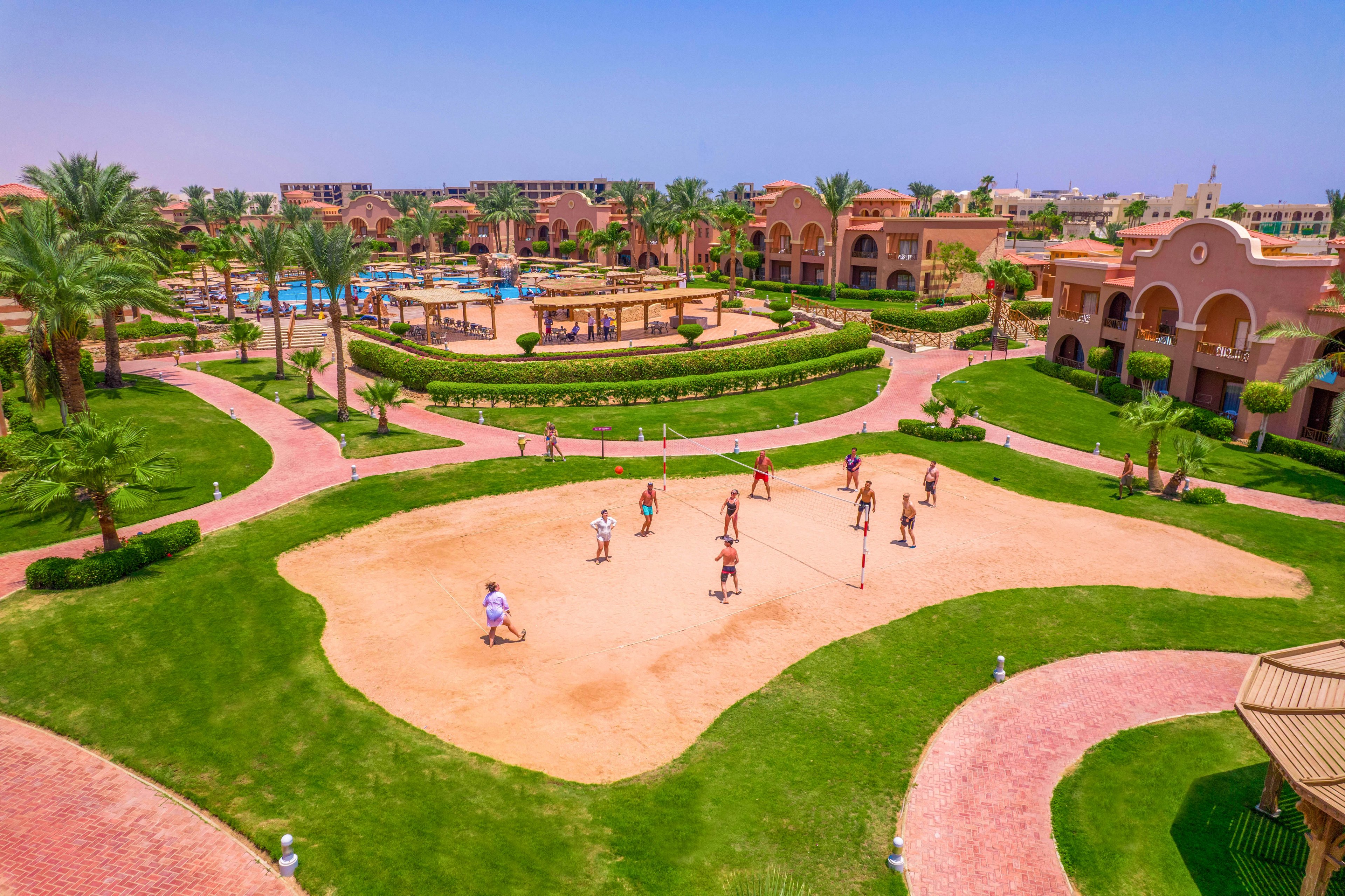 A group of people playing volleyball on a sandy court in a resort setting. The area is surrounded by vibrant greenery, palm trees, and well-maintained pathways. In the background, there are pools, lounge areas, and Mediterranean-style buildings under a clear blue sky.