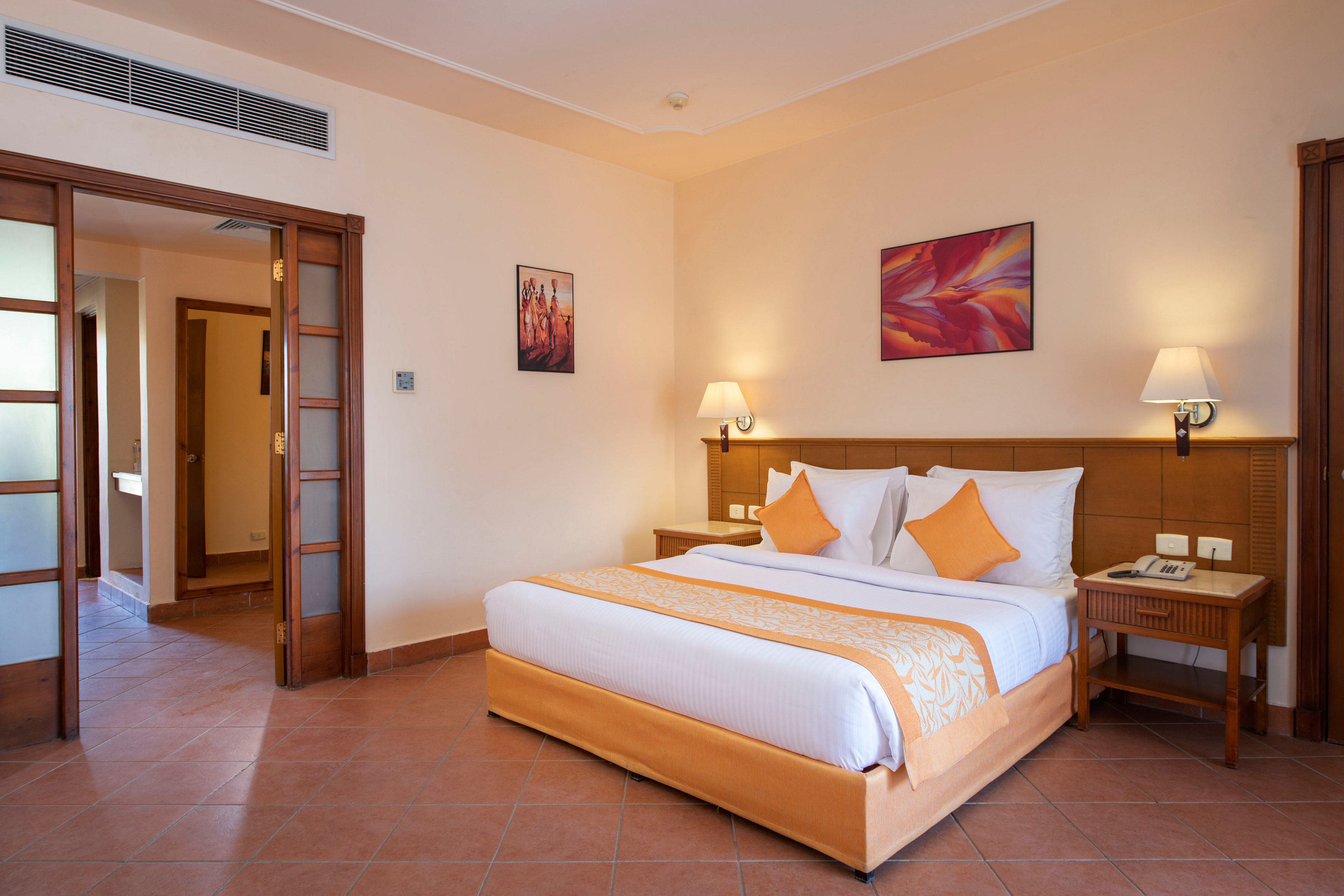 A well-lit hotel room featuring a neatly made double bed with orange accents and matching pillows. The room includes bedside tables, wall-mounted lamps, and framed artwork on the walls. A wooden sliding door leads to an adjacent area, and the tiled flooring complements the warm tones of the decor.