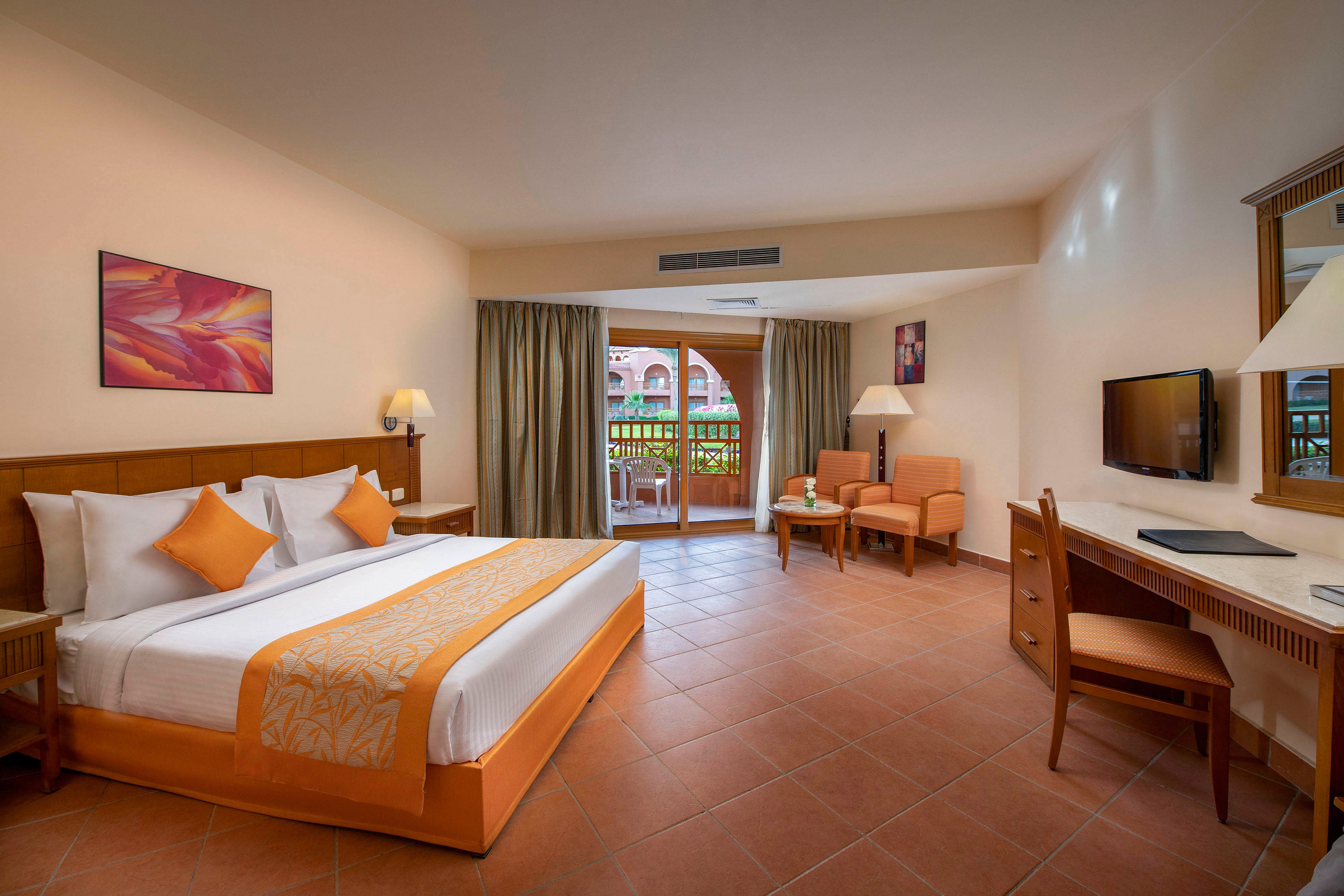 A spacious hotel room featuring a large bed with orange accents, a seating area, and a desk. The room includes a flat-screen TV, warm lighting, and a balcony with a view of greenery. The decor is modern and inviting, with earthy tones and tiled flooring.