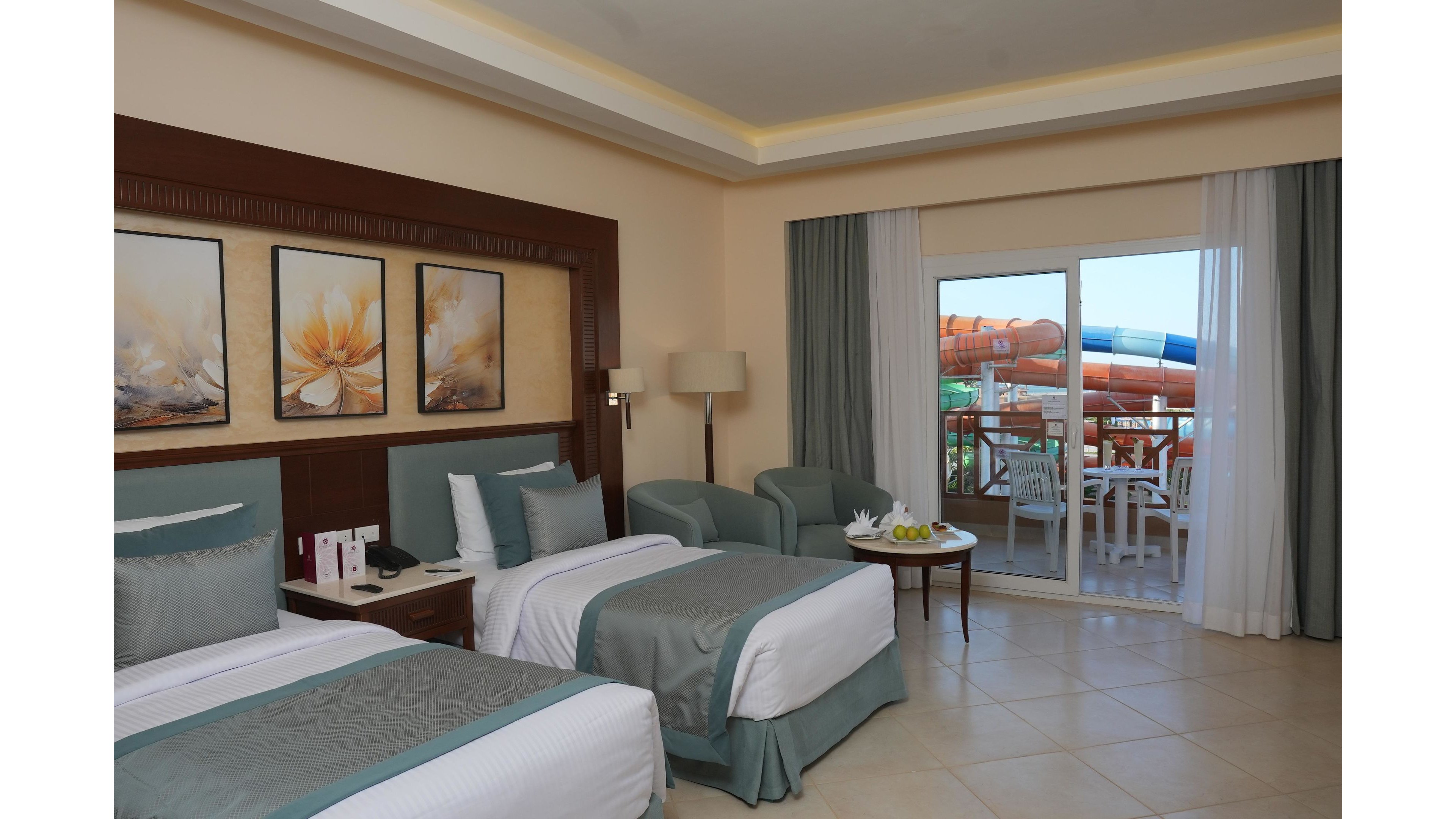 A contemporary hotel room features two neatly made twin beds with soft blue and white bedding. Comfortable armchairs and a small round table with fruit sit near a large glass door. Outside, a balcony with white chairs and table overlooks bright, twisting waterslides. Warm lighting, tiled floors, and floral artwork create a clean, inviting atmosphere.