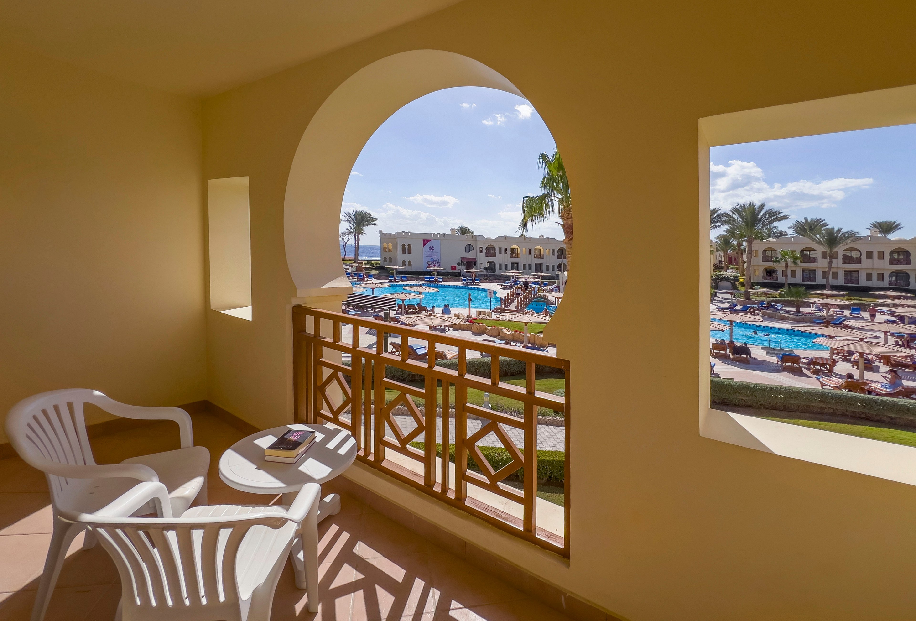 A scenic view from a resort balcony featuring a pool area surrounded by palm trees and lounge chairs. The balcony includes two white plastic chairs and a small round table, creating a relaxing outdoor seating space. The bright, sunny weather enhances the inviting atmosphere of the resort.