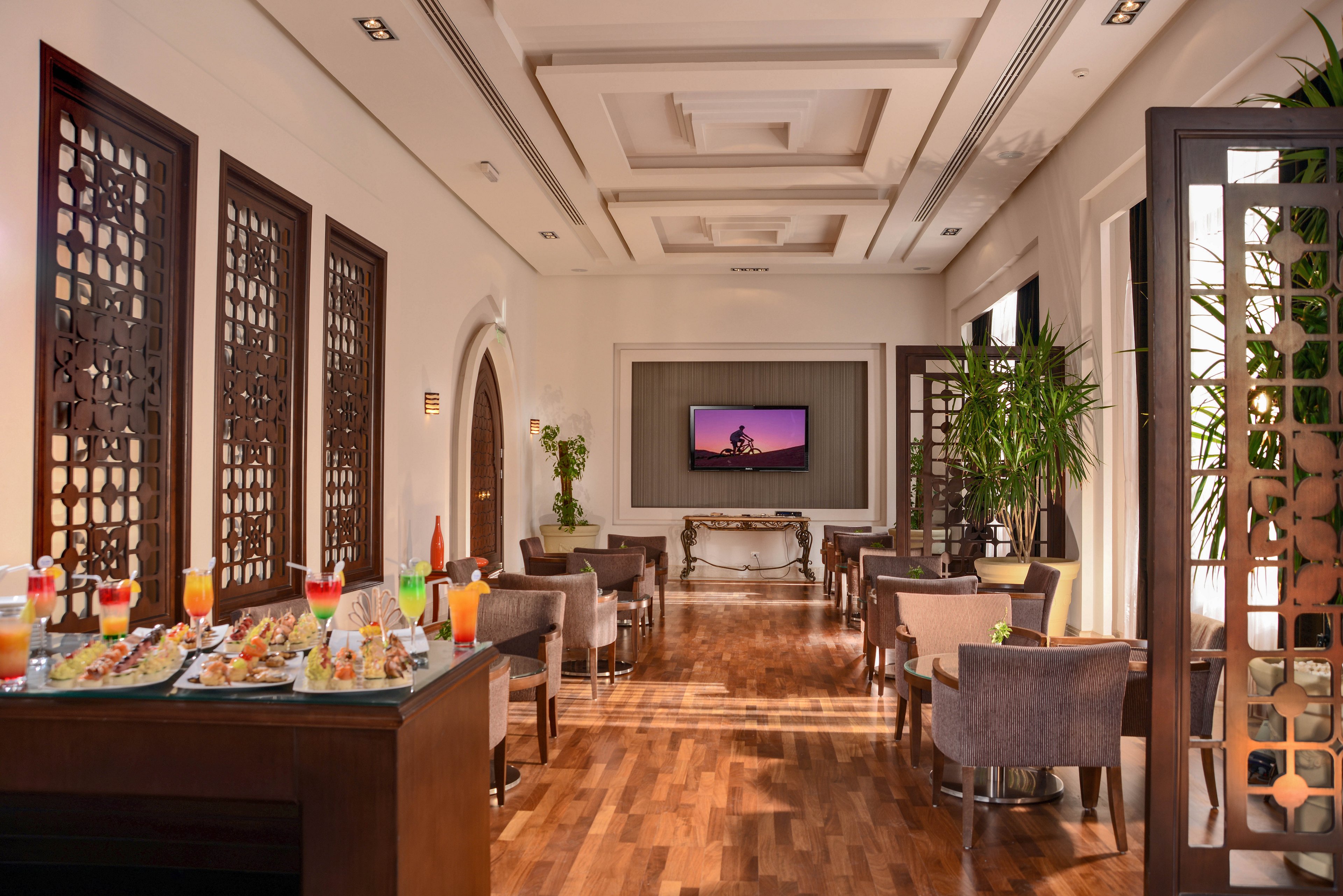 A sophisticated lounge area featuring wooden flooring, modern decor, and intricate panel designs. A buffet table is set with colorful beverages and snacks, while comfortable seating is arranged throughout the space. A wall-mounted screen displays a scenic image, adding to the ambiance.