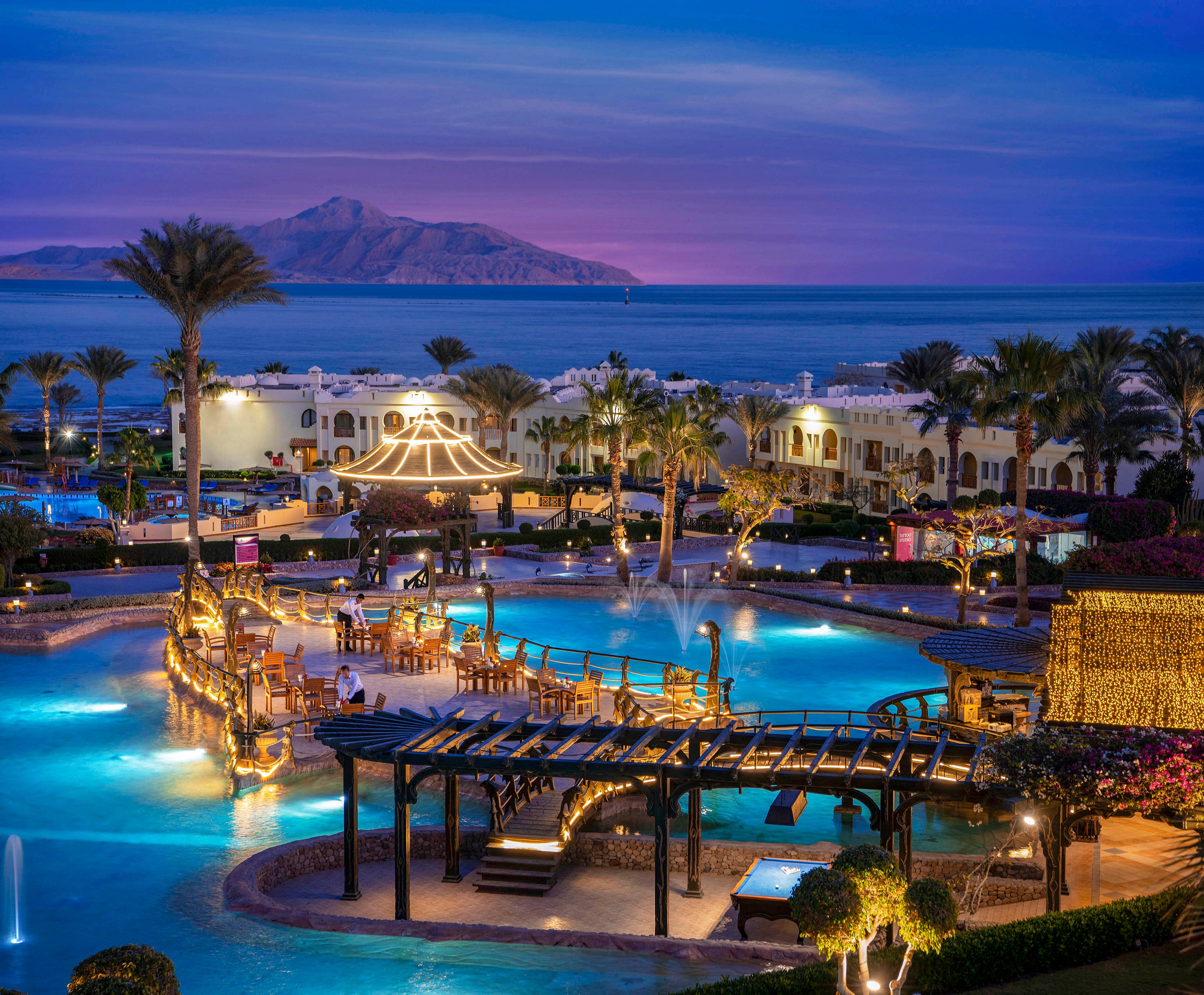 A stunning resort pool illuminated by warm evening lights, surrounded by palm trees and elegant seating areas. The scene overlooks a serene ocean with a mountain in the distance under a twilight sky. The setting exudes relaxation and luxury, perfect for a tranquil getaway.