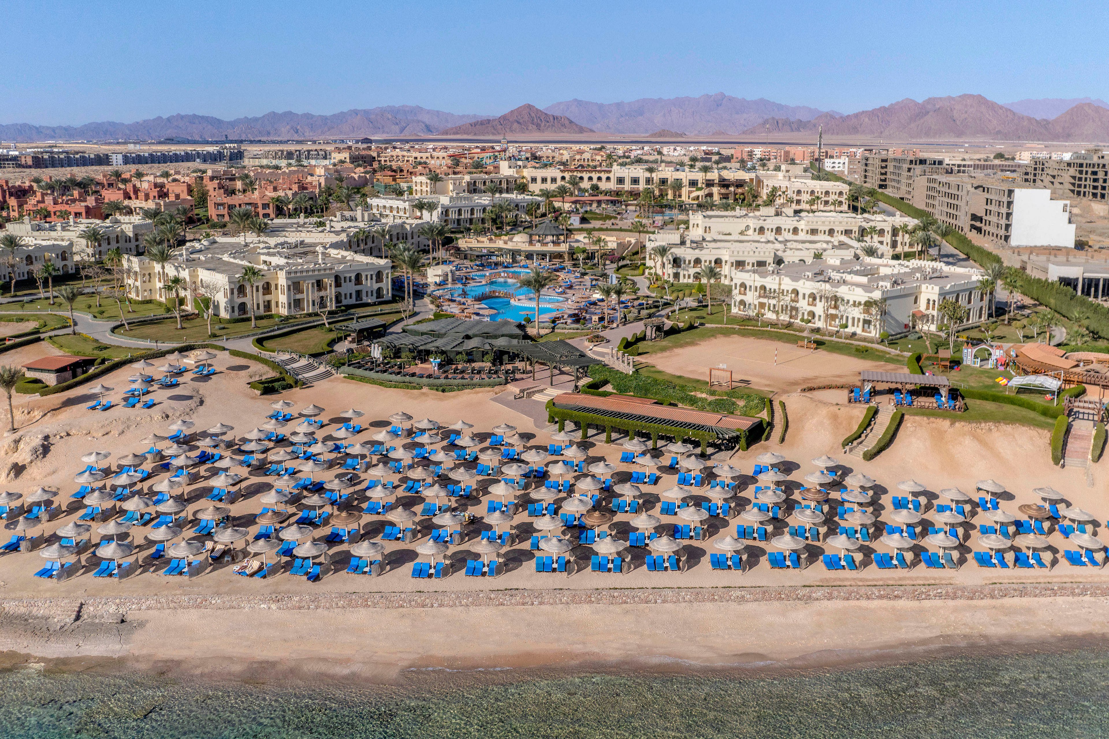 An aerial view of a luxurious beachfront resort featuring rows of blue umbrellas on a sandy beach. The resort includes multiple buildings, lush green landscaping, and a central pool area. The clear turquoise water of the sea is visible in the foreground, with distant mountains in the background. The setting is sunny and vibrant, ideal for a tropical getaway.