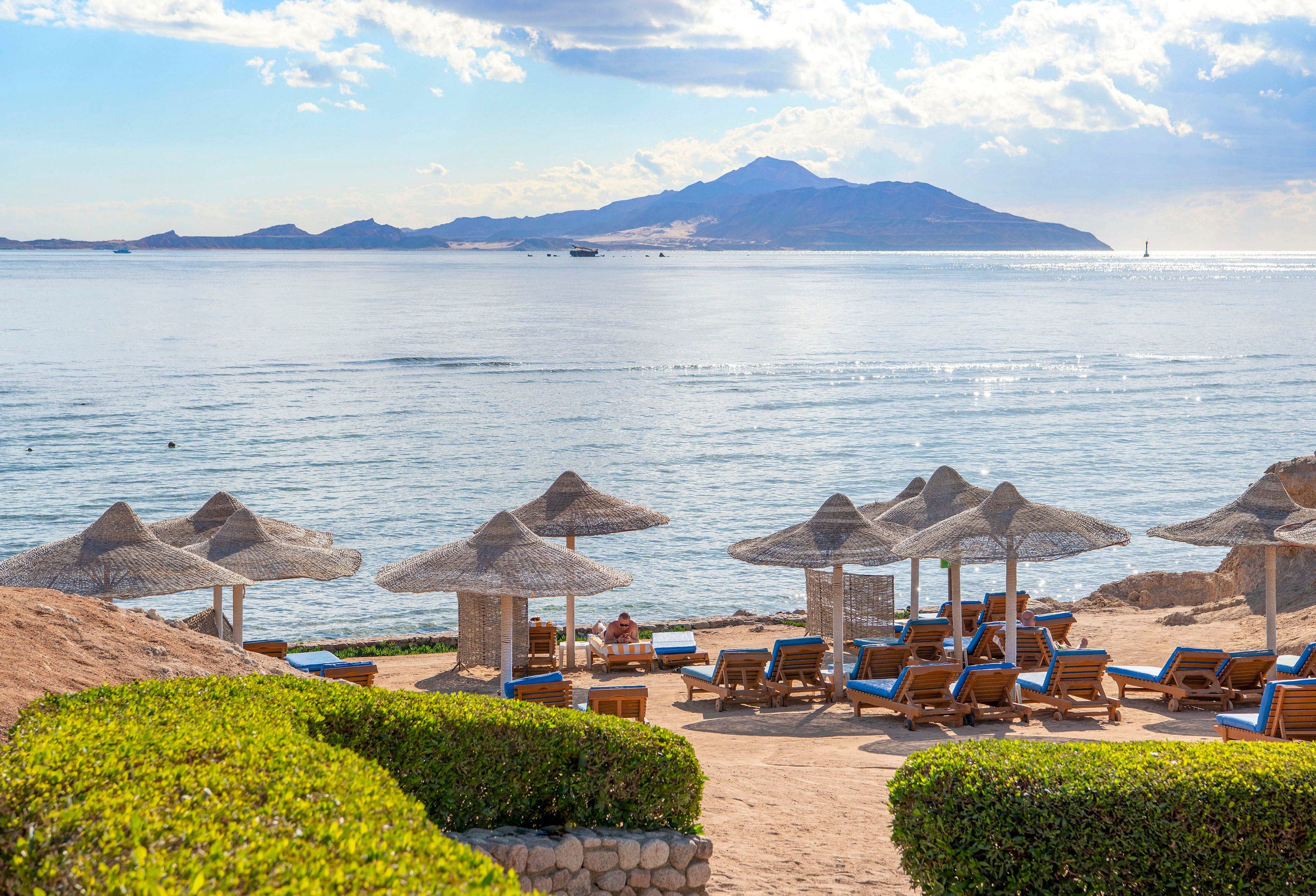 A serene beachfront setting featuring rows of straw umbrellas and wooden lounge chairs overlooking calm blue waters. The backdrop includes a distant mountain range under a partly cloudy sky. The lush green hedges in the foreground add a touch of vibrancy to the tranquil scene.