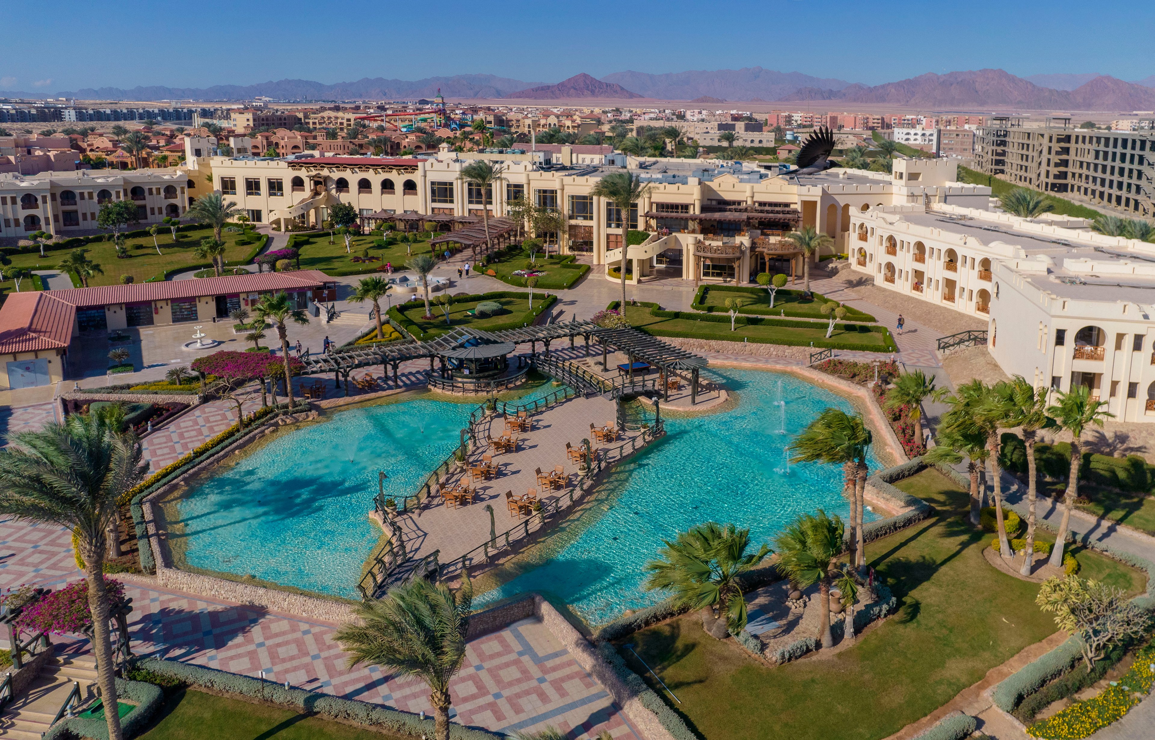 Aerial view of a luxury resort featuring a large swimming pool surrounded by lush gardens and palm trees. The resort buildings showcase elegant architecture with balconies overlooking the pool. The setting includes clear skies and distant mountains, creating a serene and inviting atmosphere.