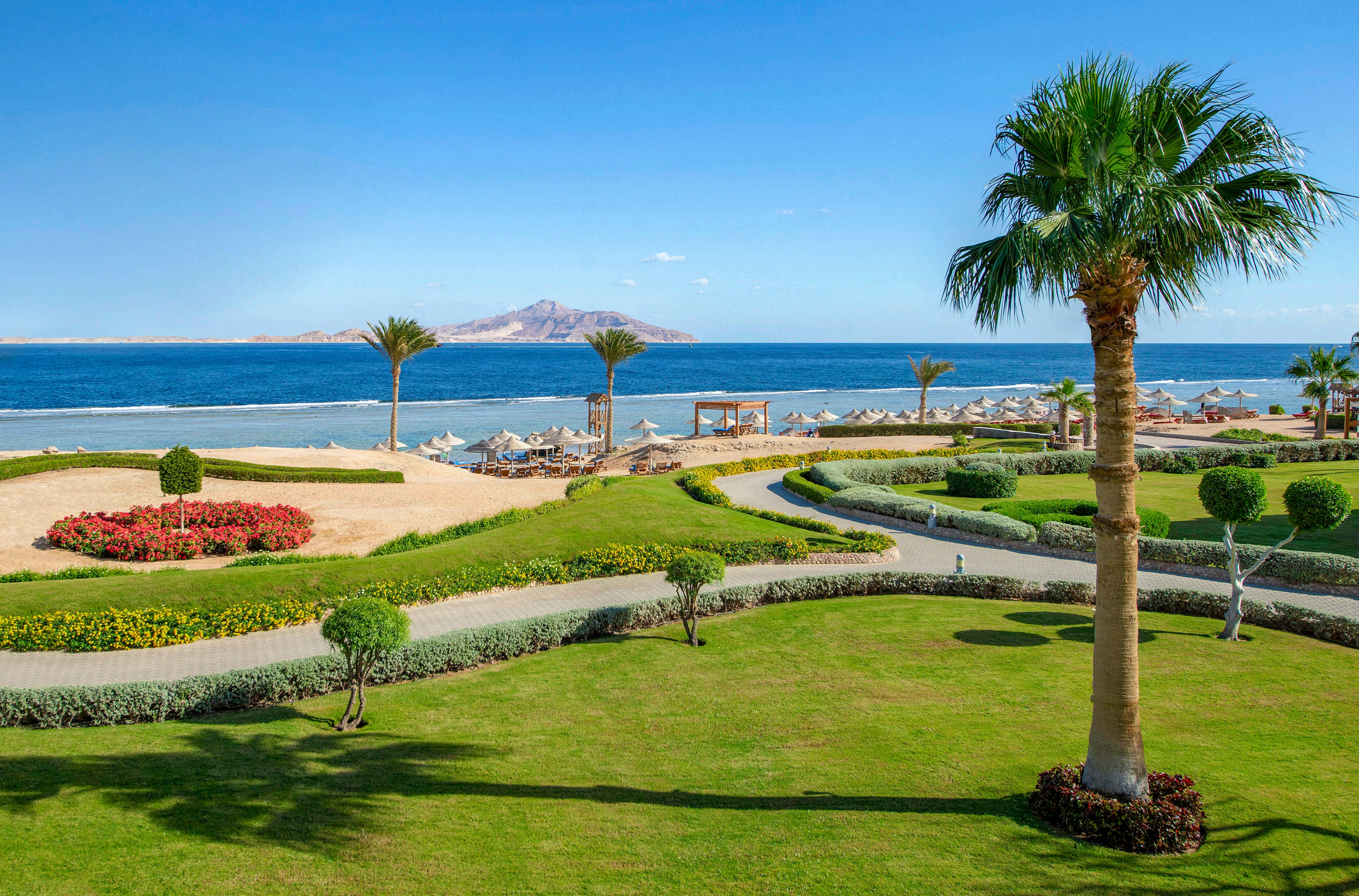 A picturesque beachfront resort featuring lush green lawns, vibrant flower beds, and tall palm trees. The setting overlooks a serene blue sea with distant mountains on the horizon. Lounge chairs and umbrellas are arranged near the shore, creating a relaxing vacation atmosphere.