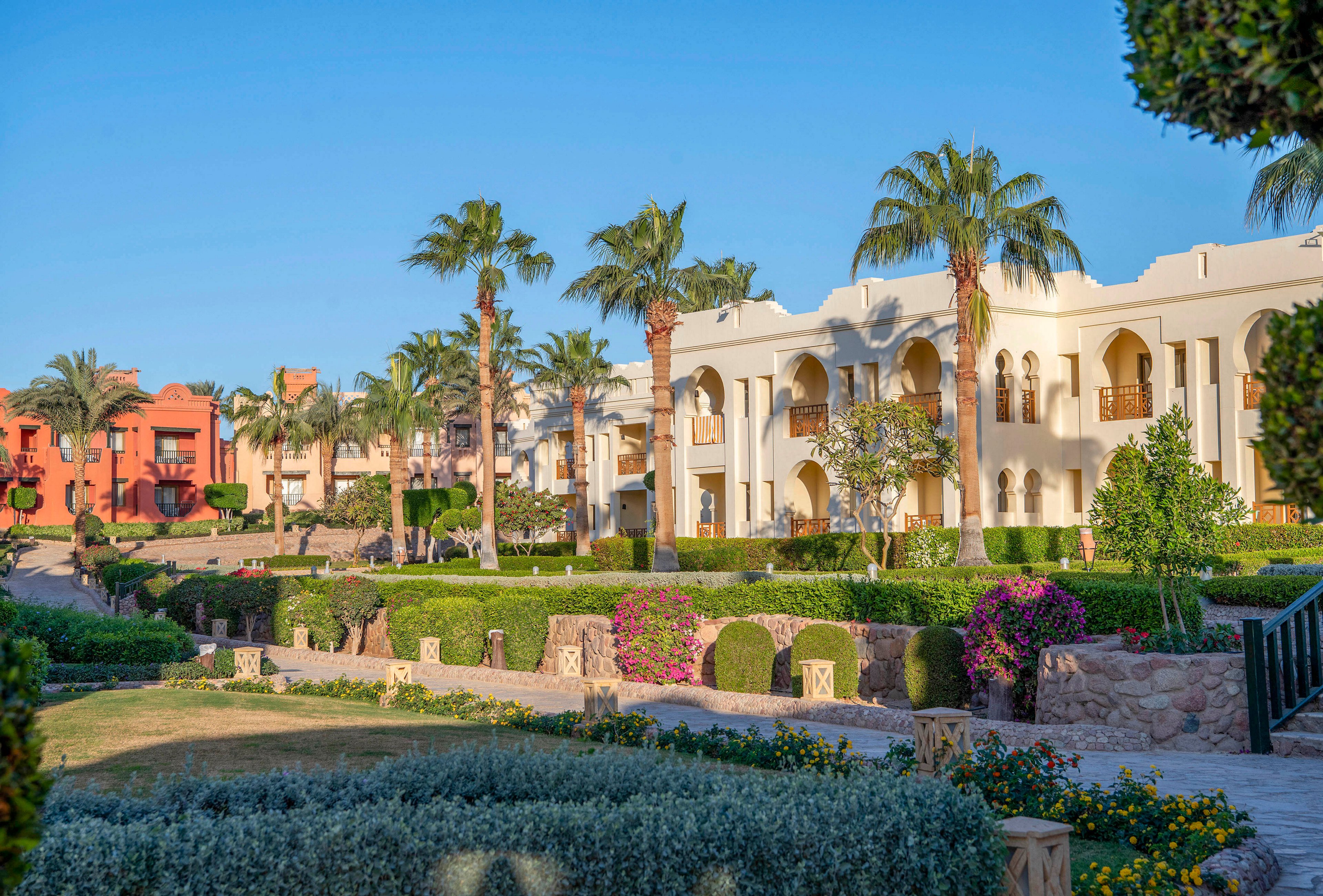 A scenic view of a Mediterranean-style resort featuring white and terracotta buildings surrounded by vibrant greenery and palm trees. The landscaped gardens include colorful flowers and neatly trimmed hedges. The setting is bright and sunny, evoking a tranquil and luxurious atmosphere.