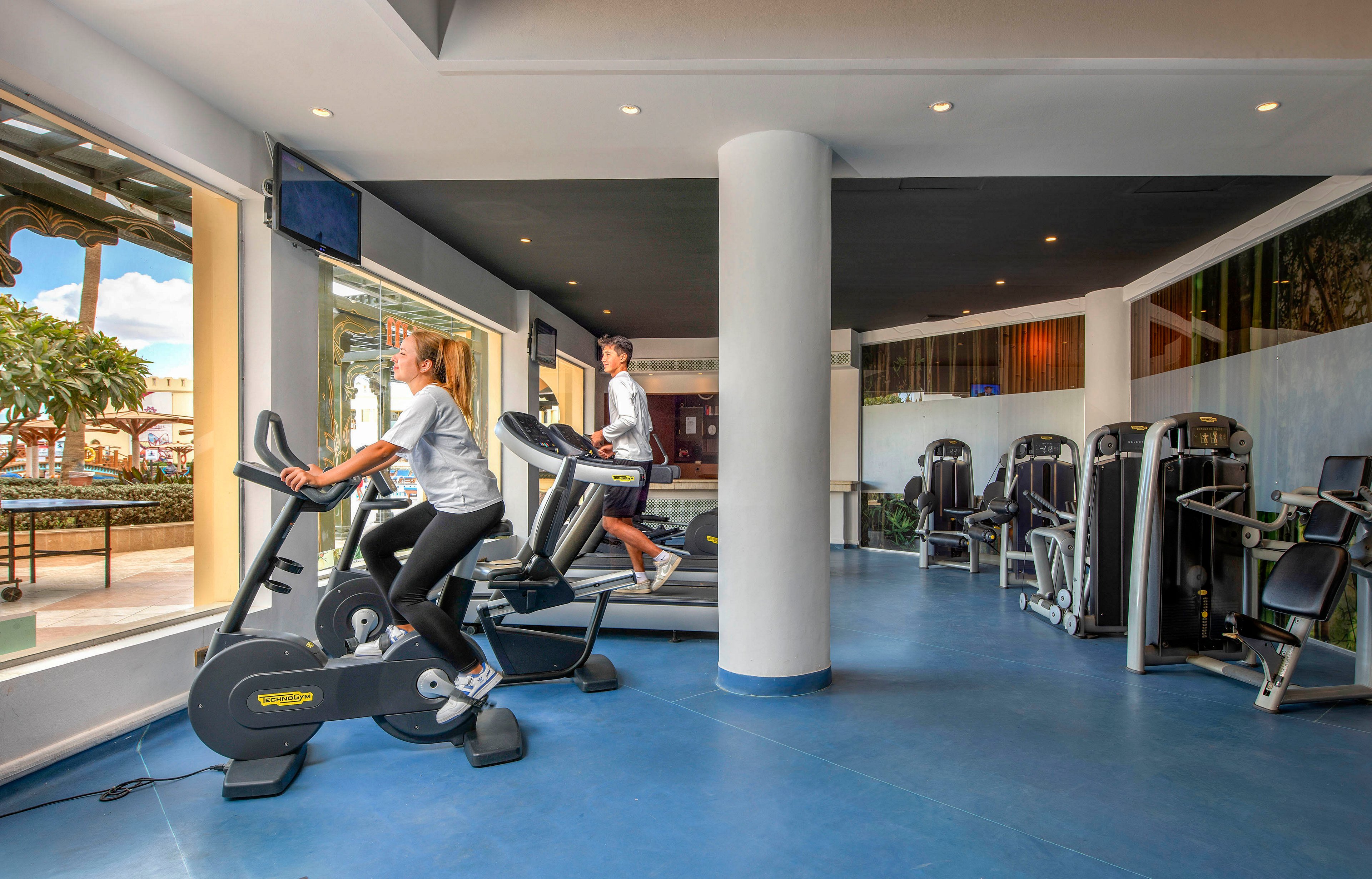 A bright and spacious gym featuring exercise bikes and other fitness equipment. Two individuals, a man and a woman, are actively using the bikes near large windows with outdoor views. The setting is clean and modern, with a blue floor and natural light enhancing the space. The gym includes additional workout machines in the background.