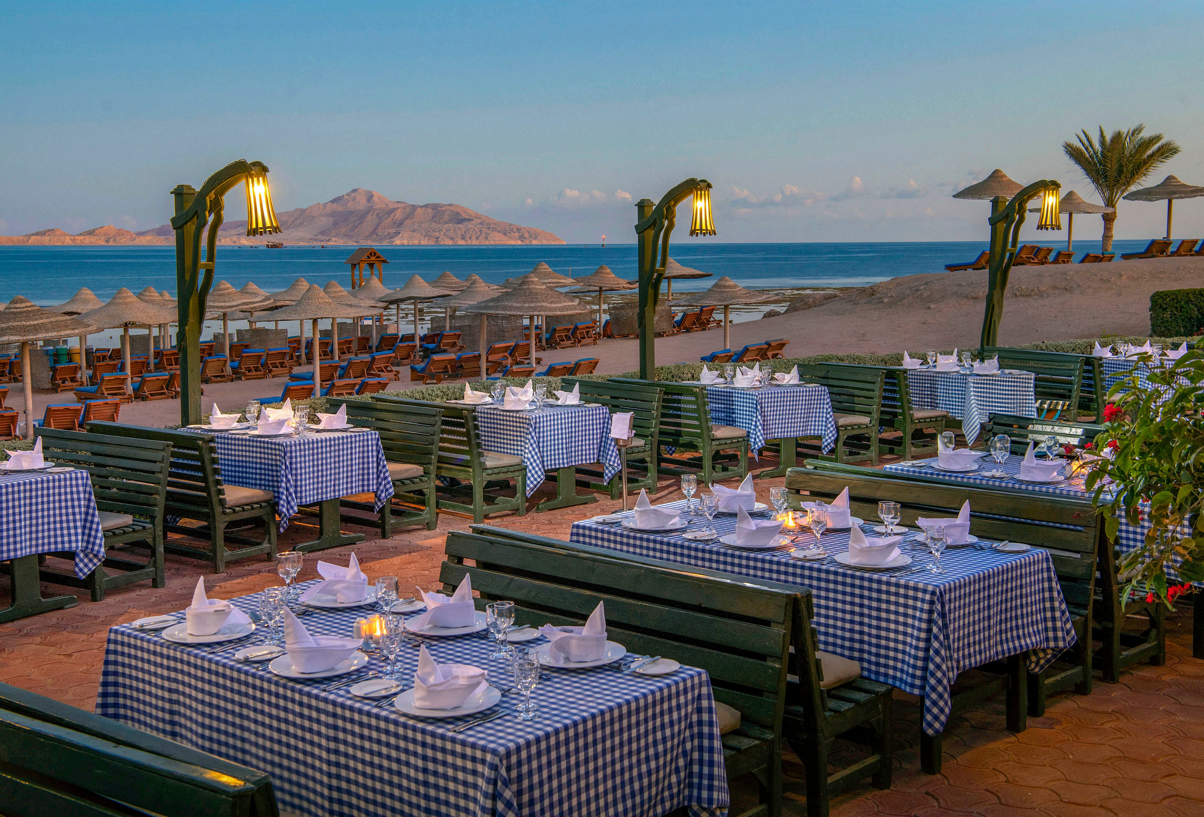 An inviting outdoor dining area with tables covered in blue and white checkered tablecloths, set against a serene seaside backdrop. The scene features neatly arranged table settings with napkins and glassware, illuminated by warm lamppost lighting. In the distance, straw umbrellas and lounge chairs line the sandy beach, with a mountain range visible across the calm sea.