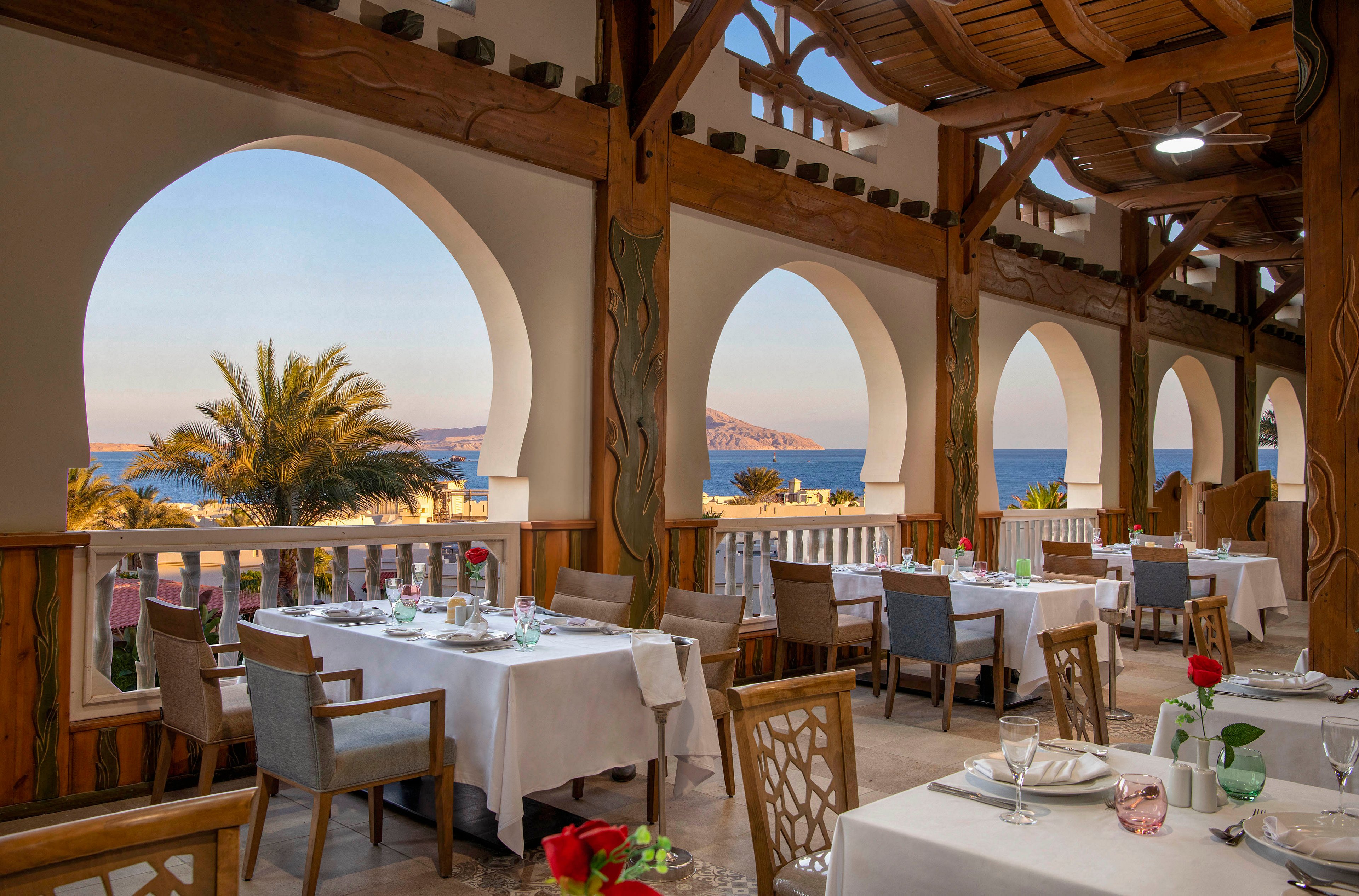 An open-air restaurant with beautifully set tables and chairs, featuring white tablecloths and red floral accents. The setting overlooks a stunning seaside view framed by arched windows and palm trees. The warm wooden decor and soft lighting create a serene and inviting atmosphere.