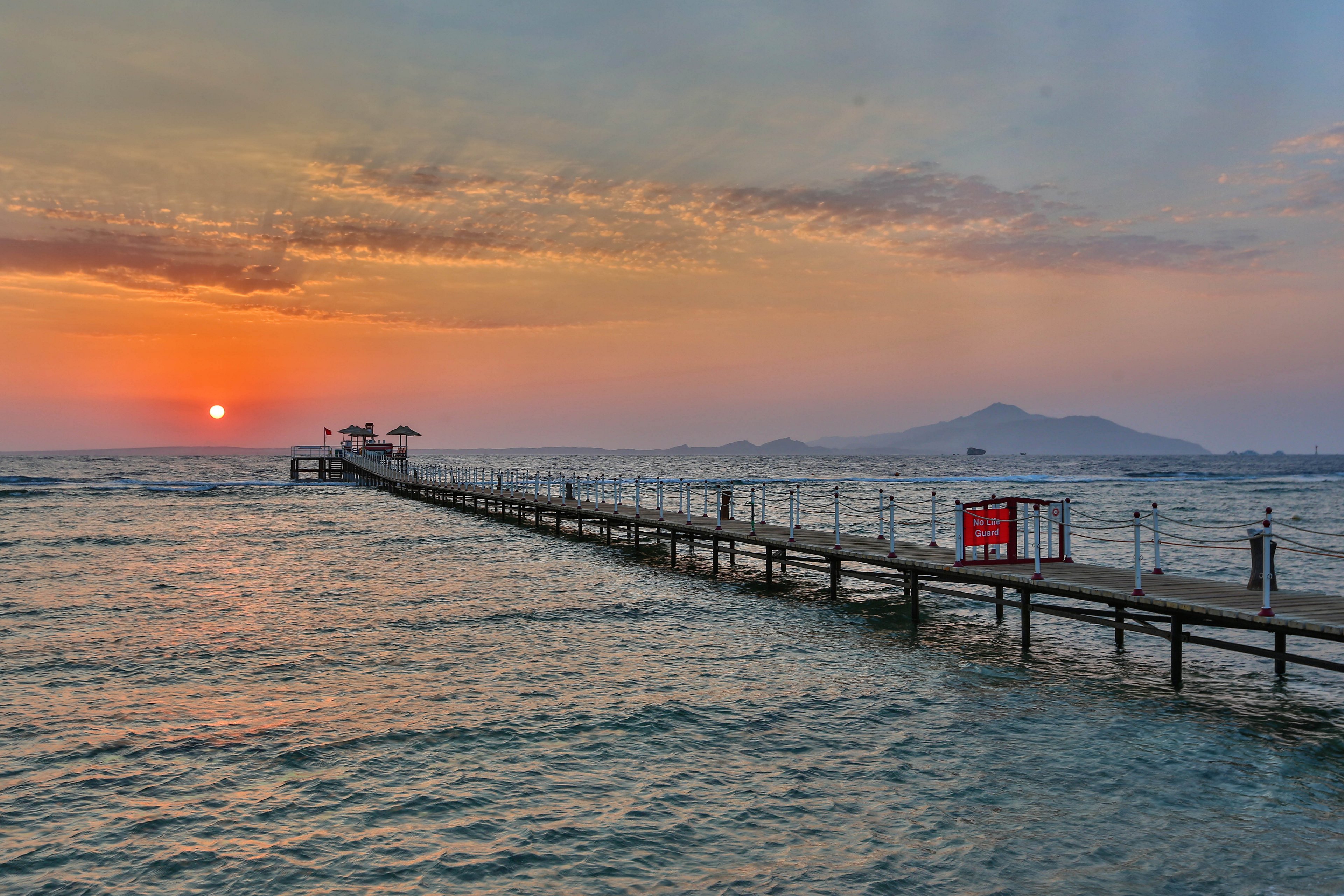 A long wooden pier stretches into the ocean, surrounded by calm waters during a vibrant sunset. The sky features warm hues of orange and pink, with scattered clouds adding texture. A red sign is visible on the pier, adding a pop of color to the serene scene. In the distance, a mountain silhouette completes the tranquil coastal view.