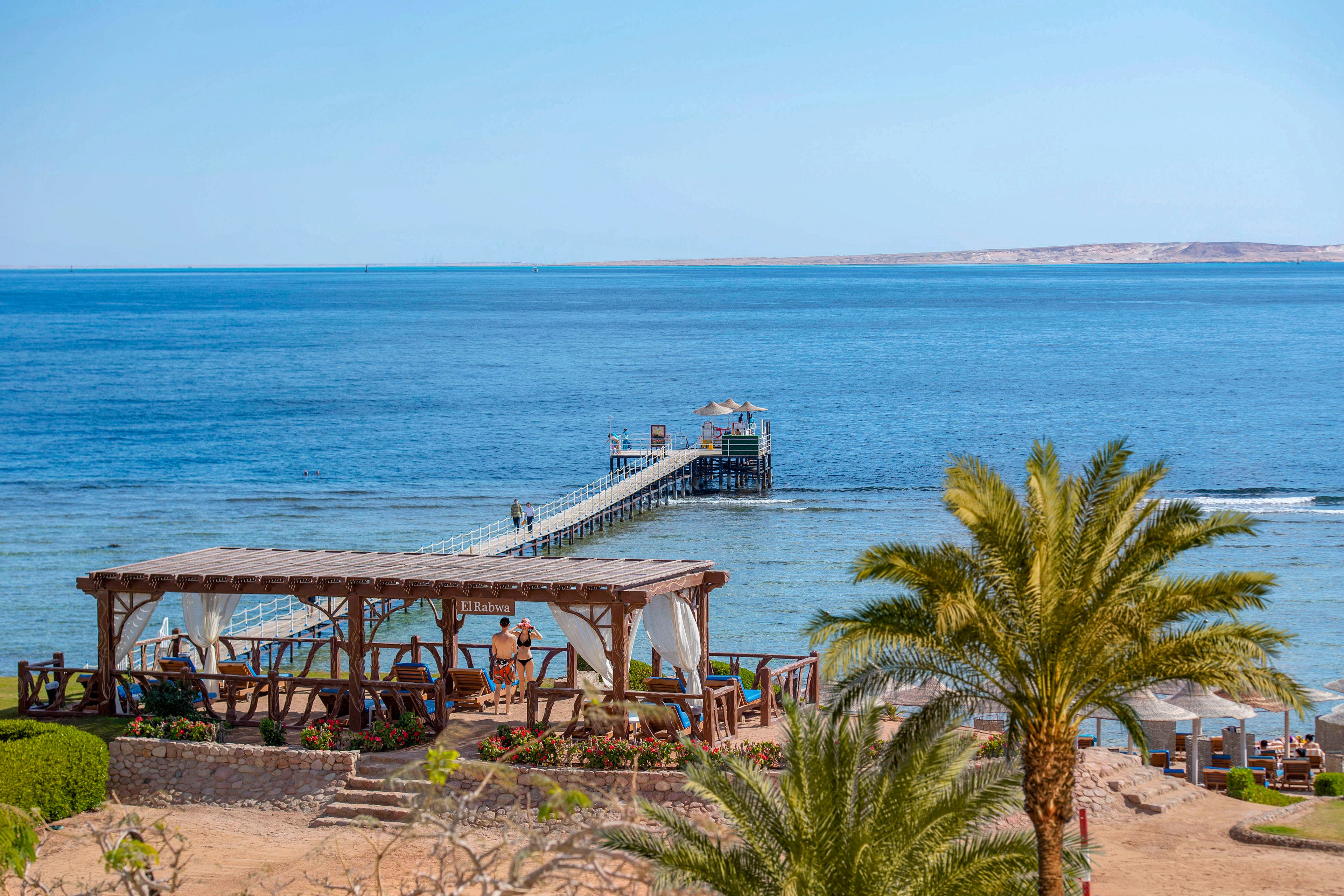 A picturesque view of a wooden pier extending into calm blue waters, surrounded by a tropical beach setting. Palm trees and shaded seating areas create a relaxing atmosphere. The clear sky and distant landmass add to the serene coastal vibe.