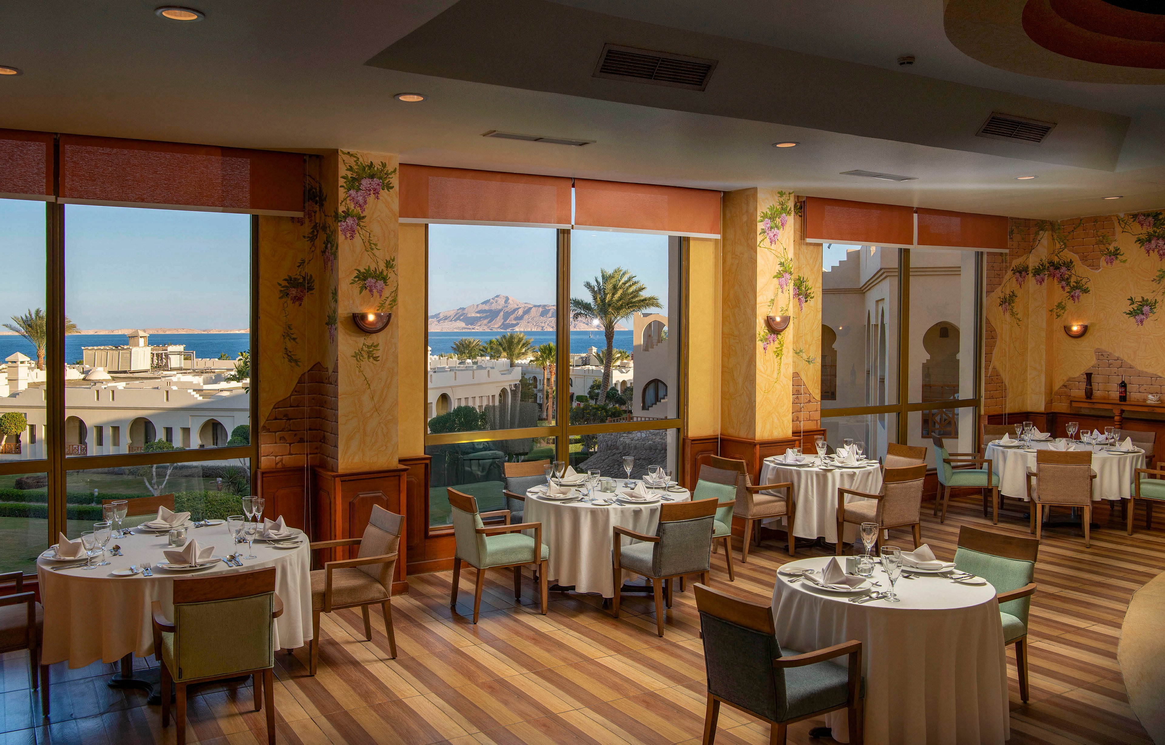 A sophisticated dining area featuring neatly arranged tables and chairs, set against large windows showcasing a stunning ocean view. The interior has warm wooden flooring and floral wall accents, creating a cozy yet upscale ambiance. The outdoor scenery includes palm trees, white buildings, and a distant mountain range under a clear sky.