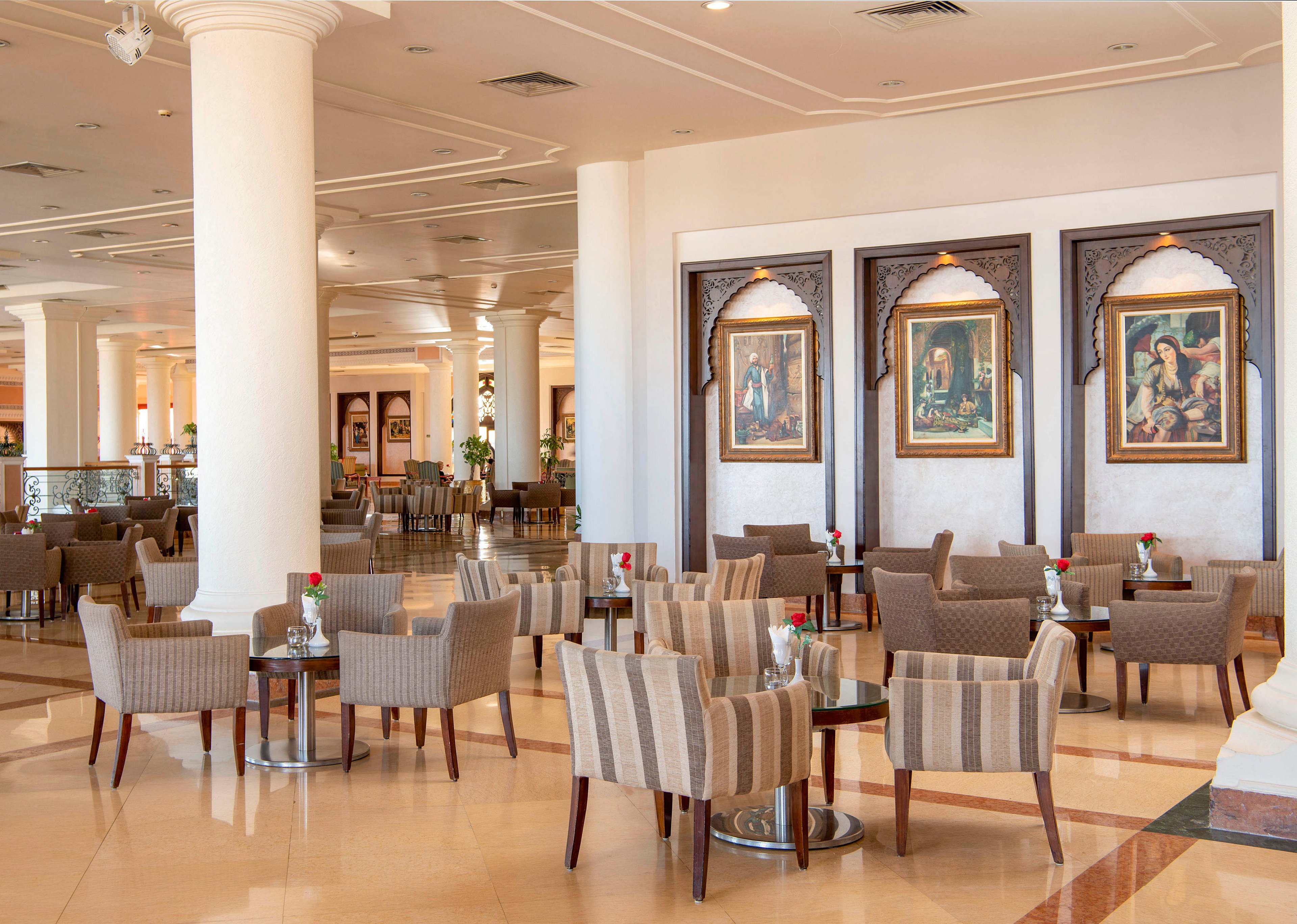 A spacious and elegant hotel lounge featuring multiple seating arrangements with striped chairs and small tables. The setting includes decorative wall art, ornate wooden frames, and polished flooring. Red roses in vases add a touch of sophistication to the ambiance.