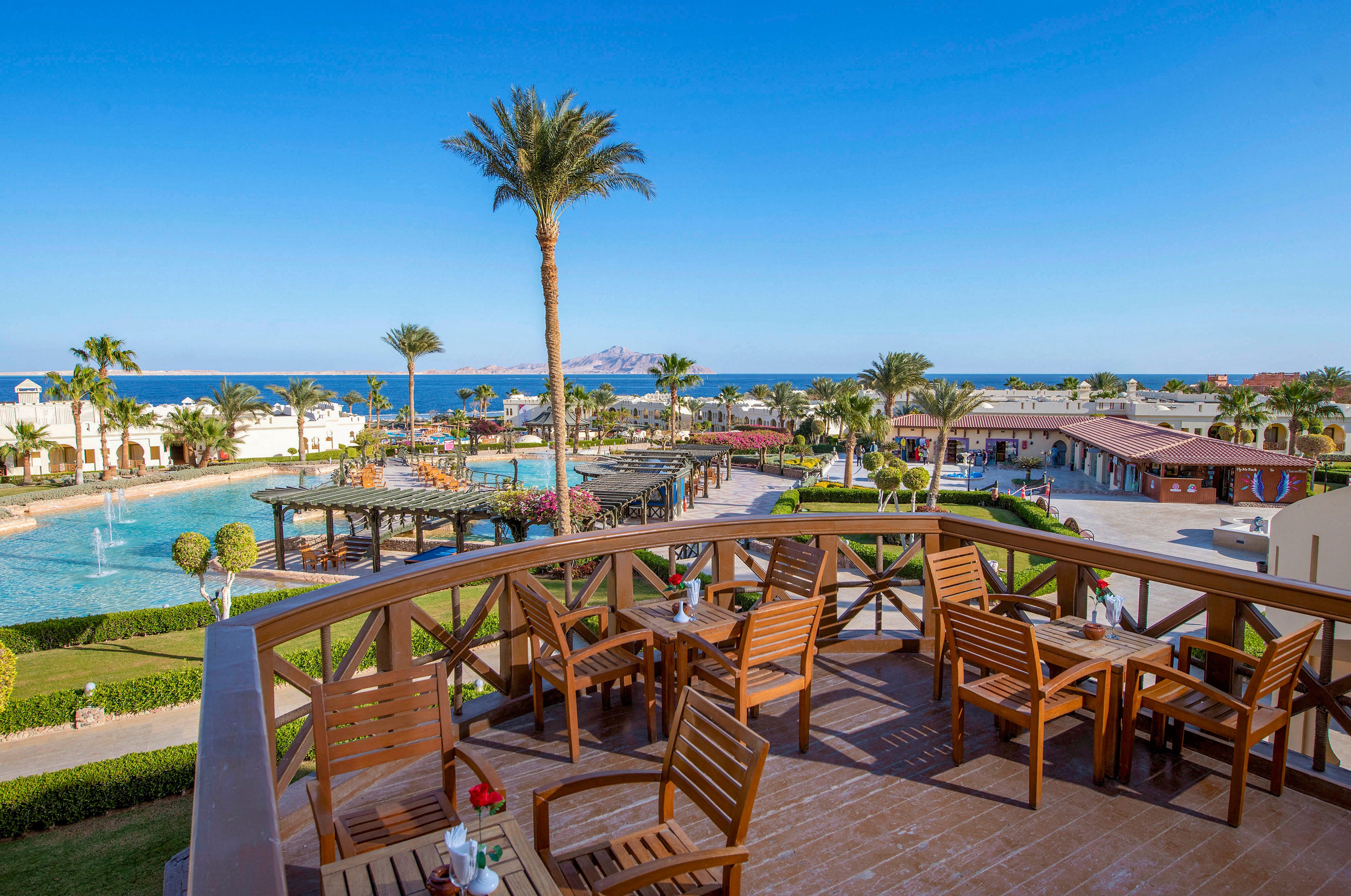 A scenic outdoor seating area with wooden chairs and tables overlooks a resort pool and the sea in the distance. The setting features lush greenery, palm trees, and a clear blue sky. The tranquil atmosphere is ideal for relaxation or dining with a view.