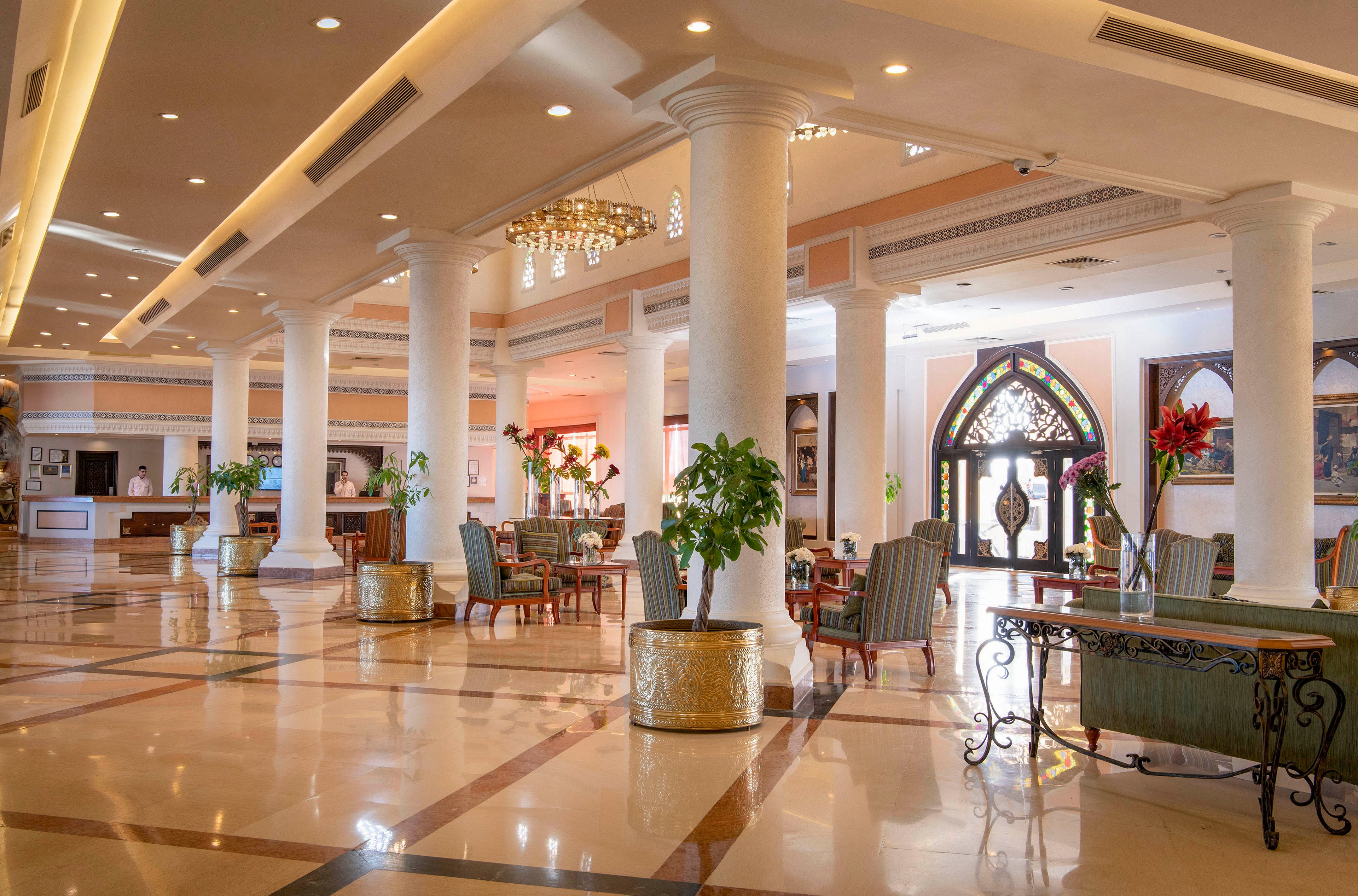 A spacious and luxurious hotel lobby featuring polished floors, tall white columns, and elegant chandeliers. Comfortable seating areas with armchairs and tables are arranged throughout the space. Large decorative plants and ornate doors enhance the sophisticated ambiance.