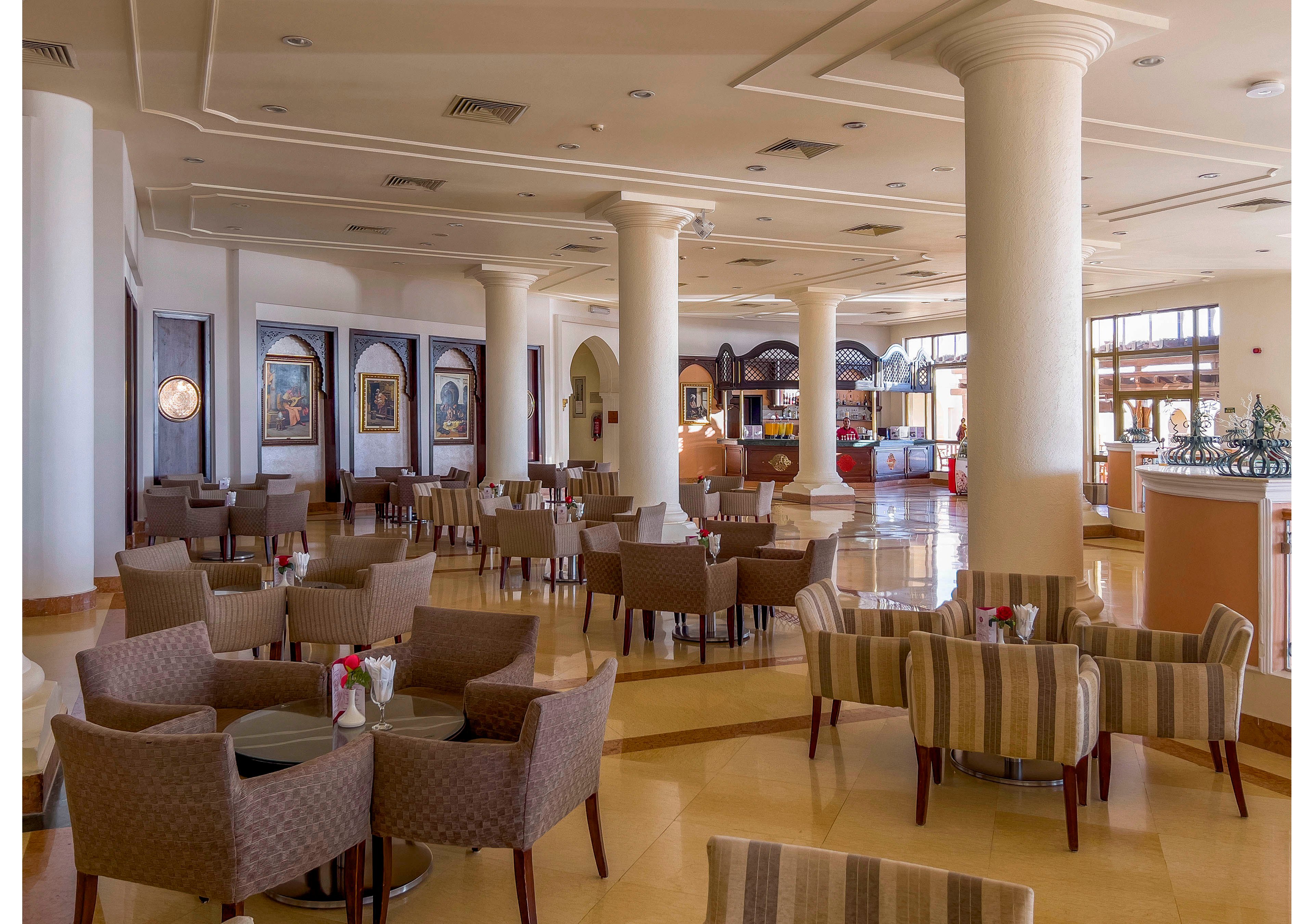 A spacious and well-lit hotel lounge featuring multiple round tables with cushioned chairs. The setting includes polished floors, white columns, and decorative wall art, creating a sophisticated ambiance. The area is designed for relaxation or casual meetings, with natural light streaming through large windows.