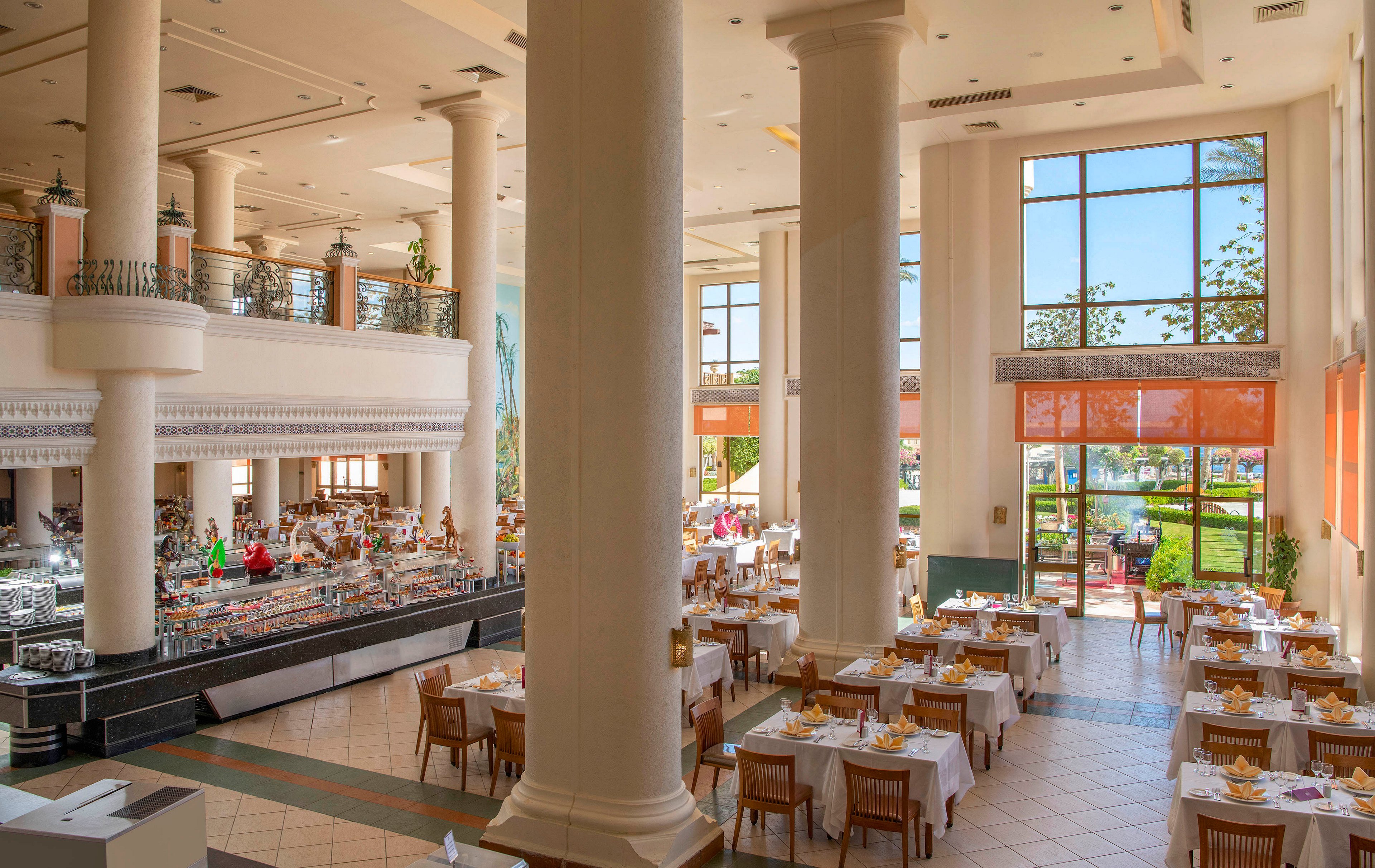 A large, well-lit restaurant featuring high ceilings, tall white columns, and neatly arranged dining tables with white tablecloths. The setting includes a buffet area and large windows offering a view of greenery and outdoor seating. The atmosphere is bright and inviting, ideal for casual or formal dining experiences.