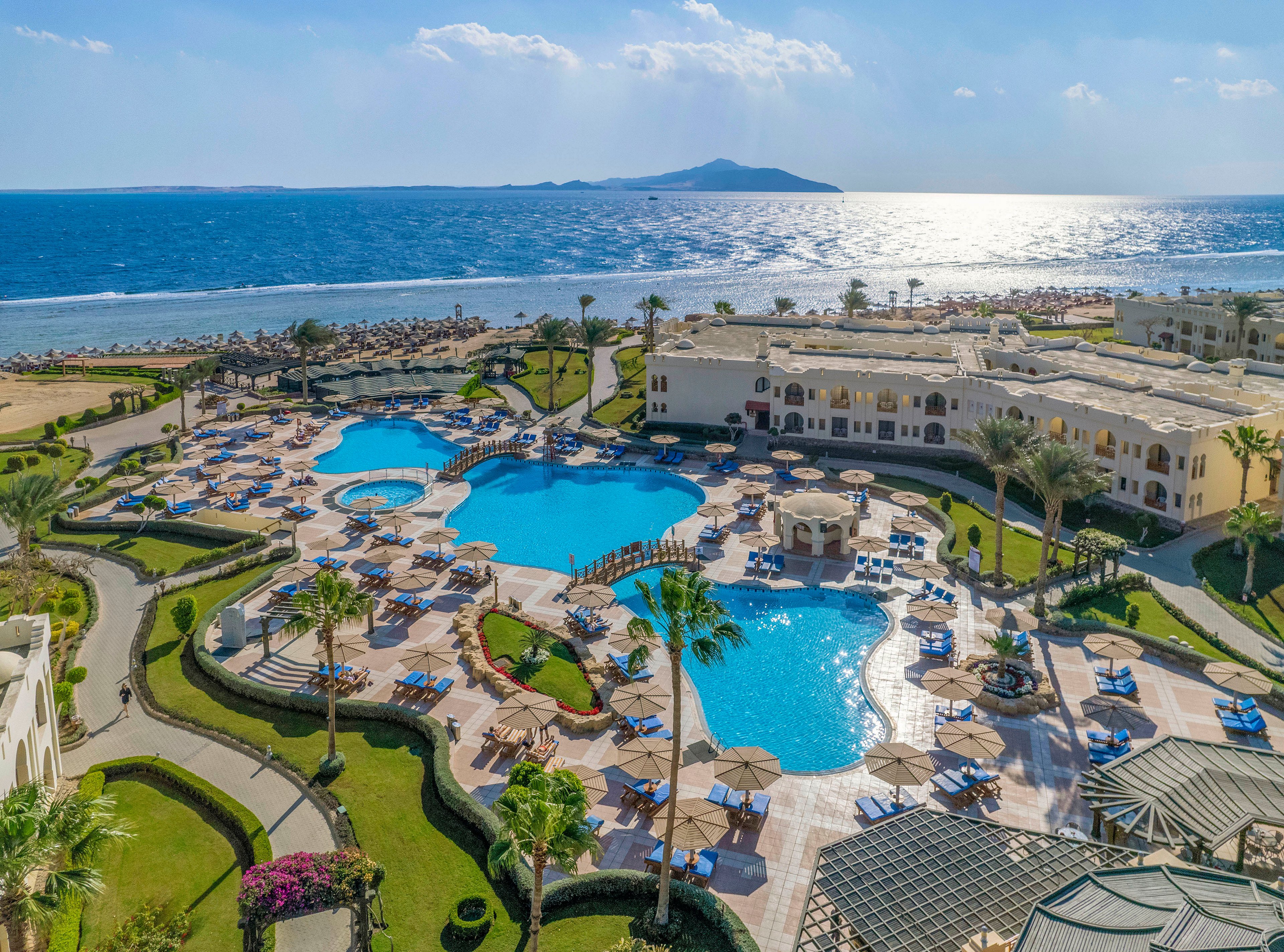 Aerial view of a luxury resort featuring multiple swimming pools, lush green landscaping, and a scenic ocean backdrop. The setting includes sun loungers, umbrellas, and well-maintained pathways. The horizon showcases a distant island under a clear blue sky.