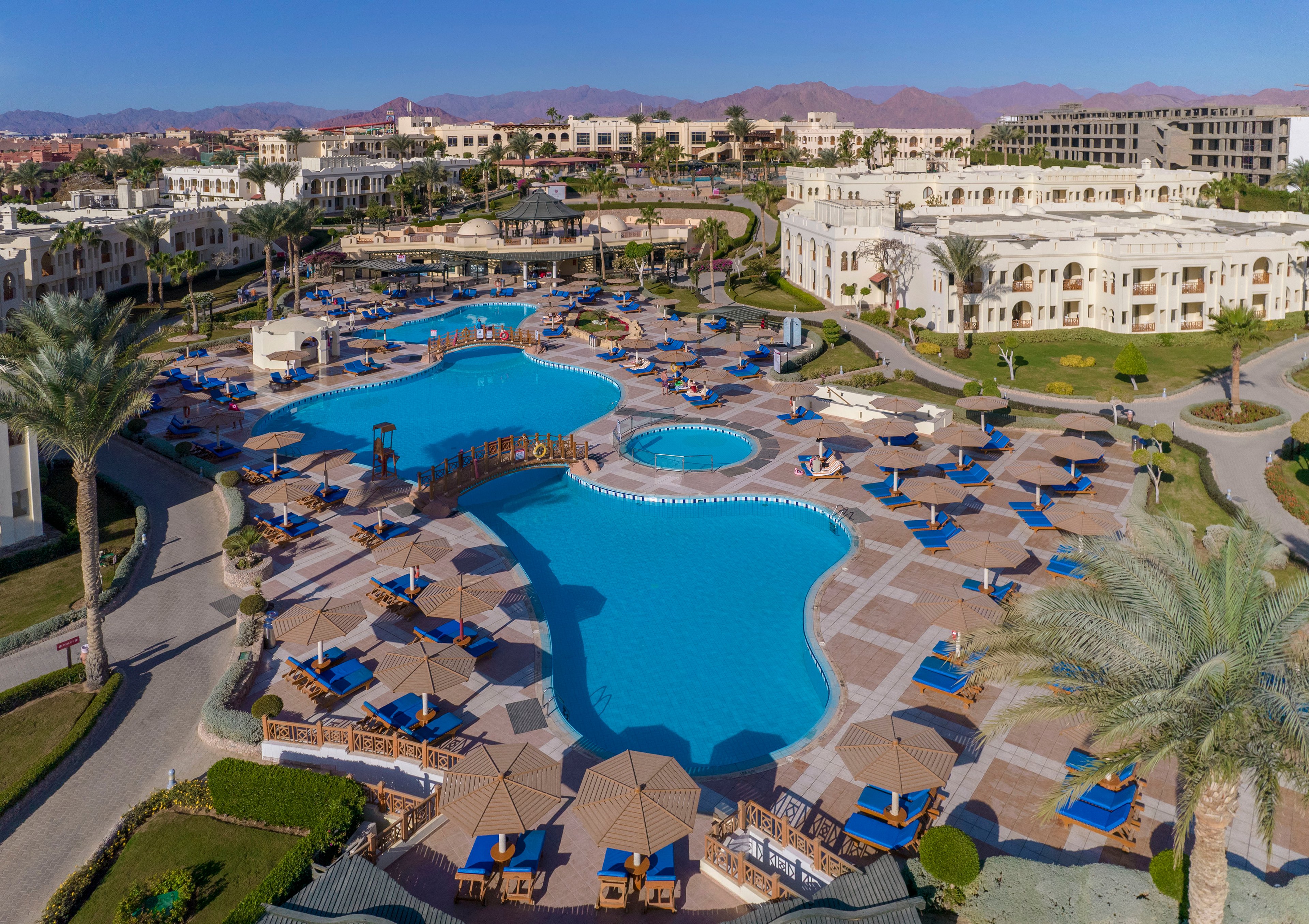 Aerial view of a luxury resort featuring multiple swimming pools surrounded by lounge chairs and umbrellas. The setting includes landscaped gardens, palm trees, and elegant architecture. The backdrop showcases mountains under a clear blue sky, creating a serene and inviting atmosphere.