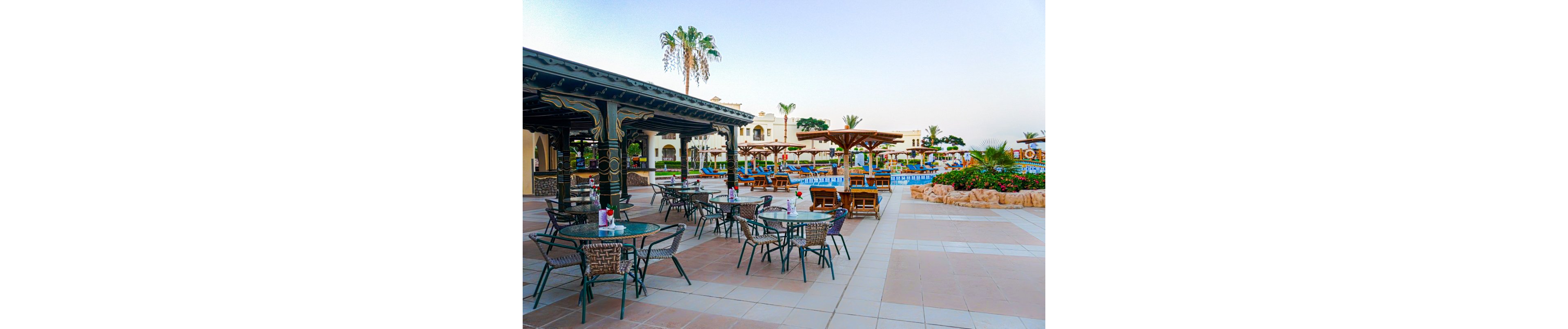 An elegant outdoor seating area with tables and chairs is set near a resort pool. The setting features palm trees, landscaped gardens, and a clear sky, creating a relaxing atmosphere. The architecture includes decorative columns and shaded areas for comfort.