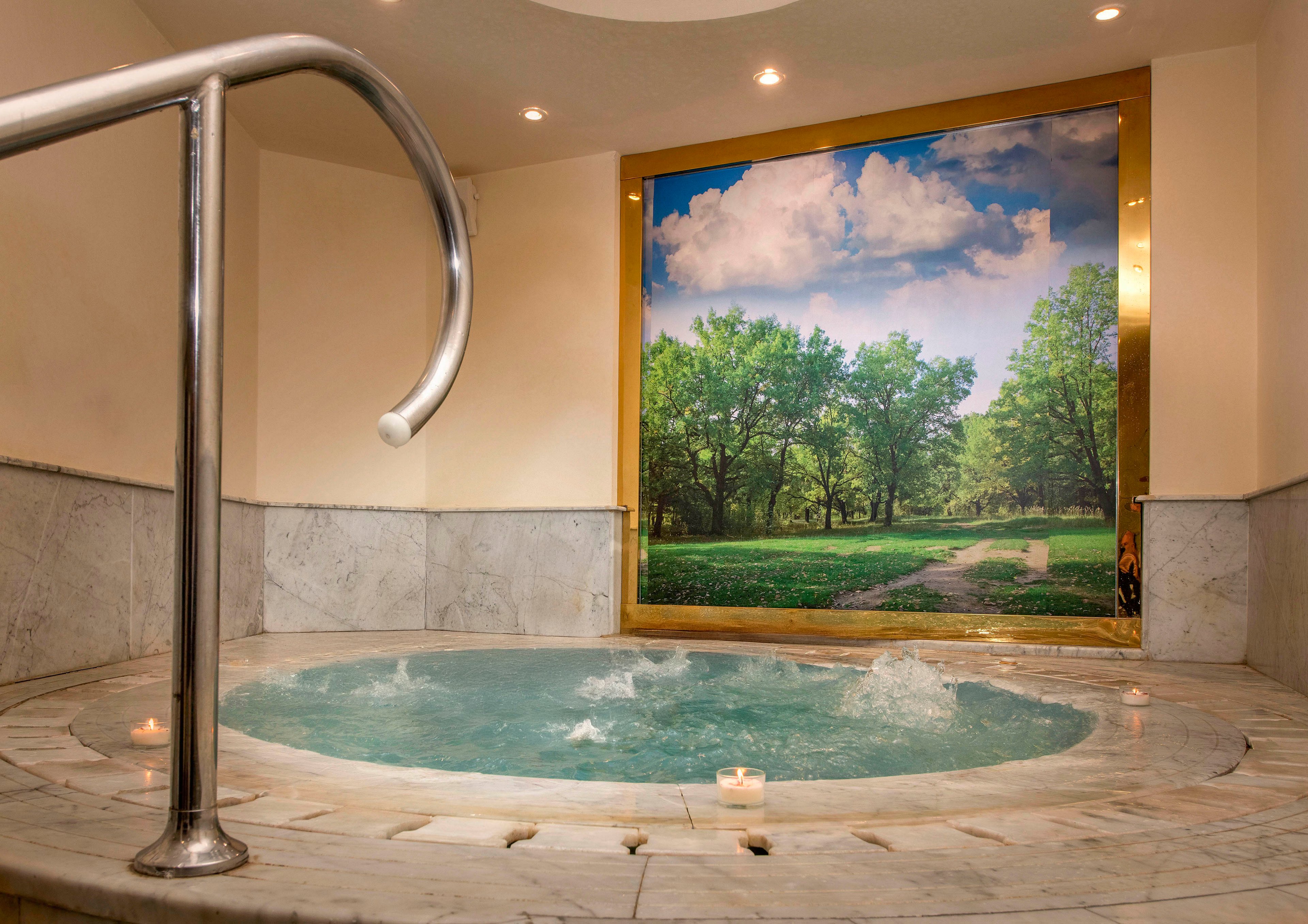 A man and woman enjoy a relaxing moment in a circular indoor hot tub surrounded by marble walls. The setting features soft lighting and a serene wall mural of a lush green park with a pathway under a blue sky. Small candles add to the tranquil ambiance.