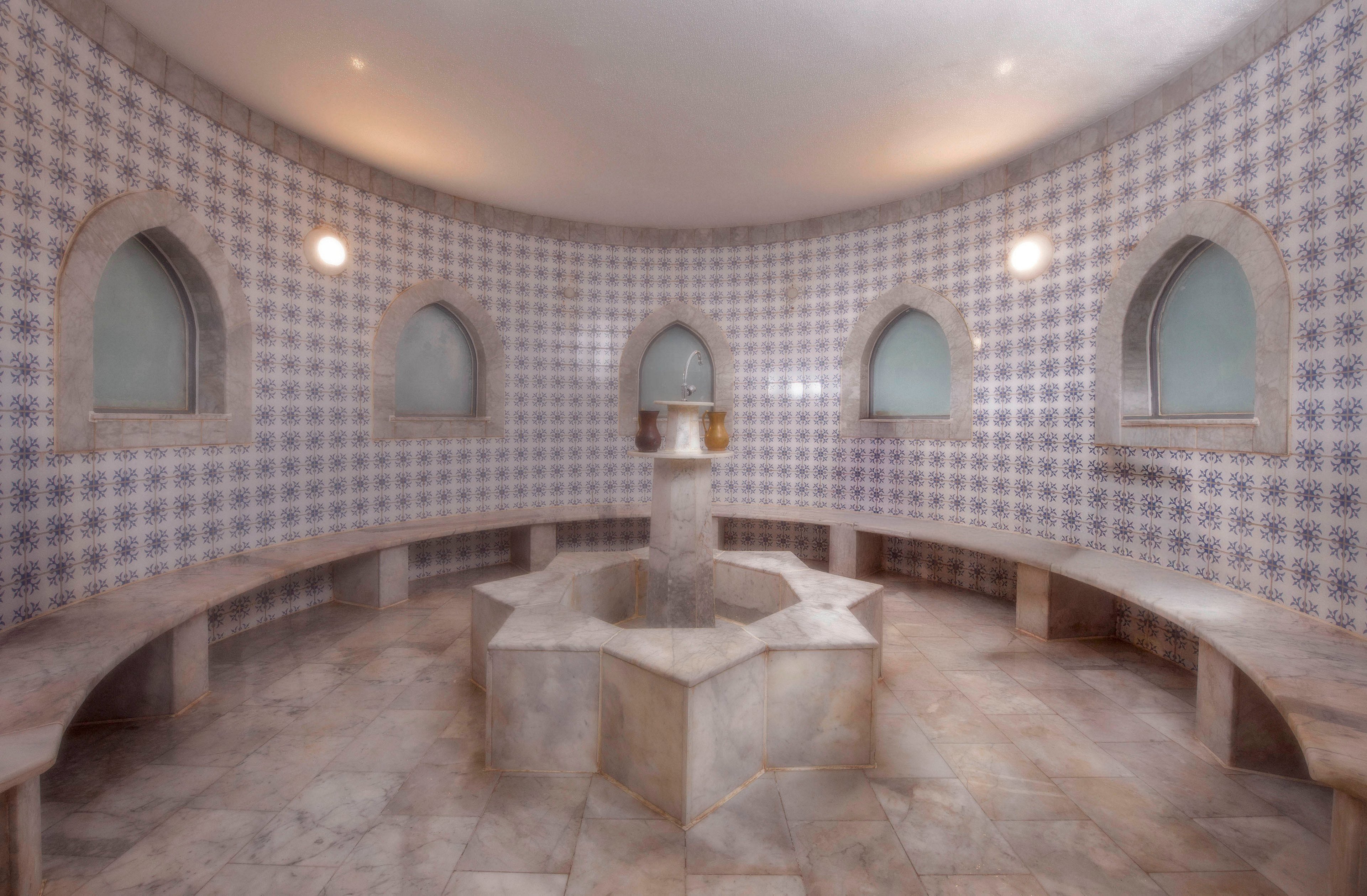 Two women are seated in a traditional tiled steam room with arched windows and a central fountain. The setting features intricate blue and white tilework, creating a serene and authentic atmosphere. The women appear relaxed, enjoying the tranquil environment of the hammam.