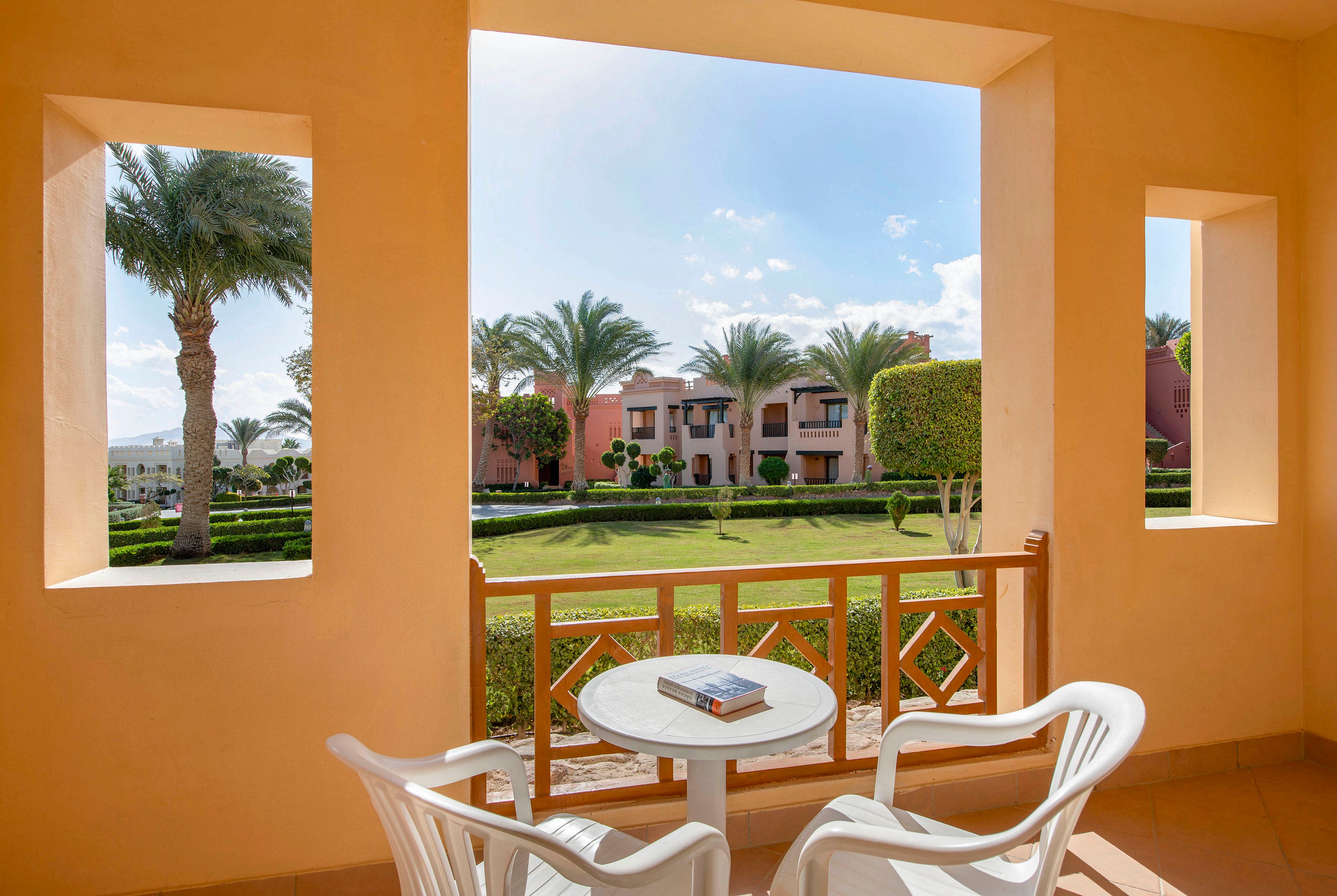A serene balcony with two white chairs and a small table overlooks a vibrant garden with palm trees and manicured lawns. The view includes colorful resort buildings under a bright blue sky. A book rests on the table, adding a touch of relaxation to the scene.